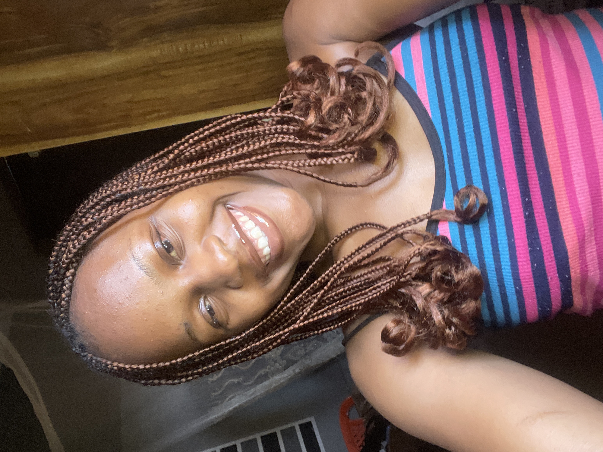 The media file is a close-up, vertically oriented selfie of a young Black woman in Jos, Nigeria. She has braided hair, styled with some braids curled at the ends. She is wearing a tank top with horizontal stripes in pink, blue, and dark blue. The woman is smiling, with her mouth open, revealing her teeth, and her eyes are open and looking towards the camera. Her skin appears to have a slight sheen, possibly from sweat or product.
The background suggests an indoor setting, with a wooden surface visible at the top of the frame. There are some dark areas in the background, possibly indicating shadows or furniture. A glimpse of a dark wall with a white horizontal railing structure can be seen in the lower part of the image. The lighting is somewhat harsh, casting shadows and highlighting the sheen on her skin. The overall impression is casual and relaxed, likely taken during daylight hours. No visible text is present in the image.