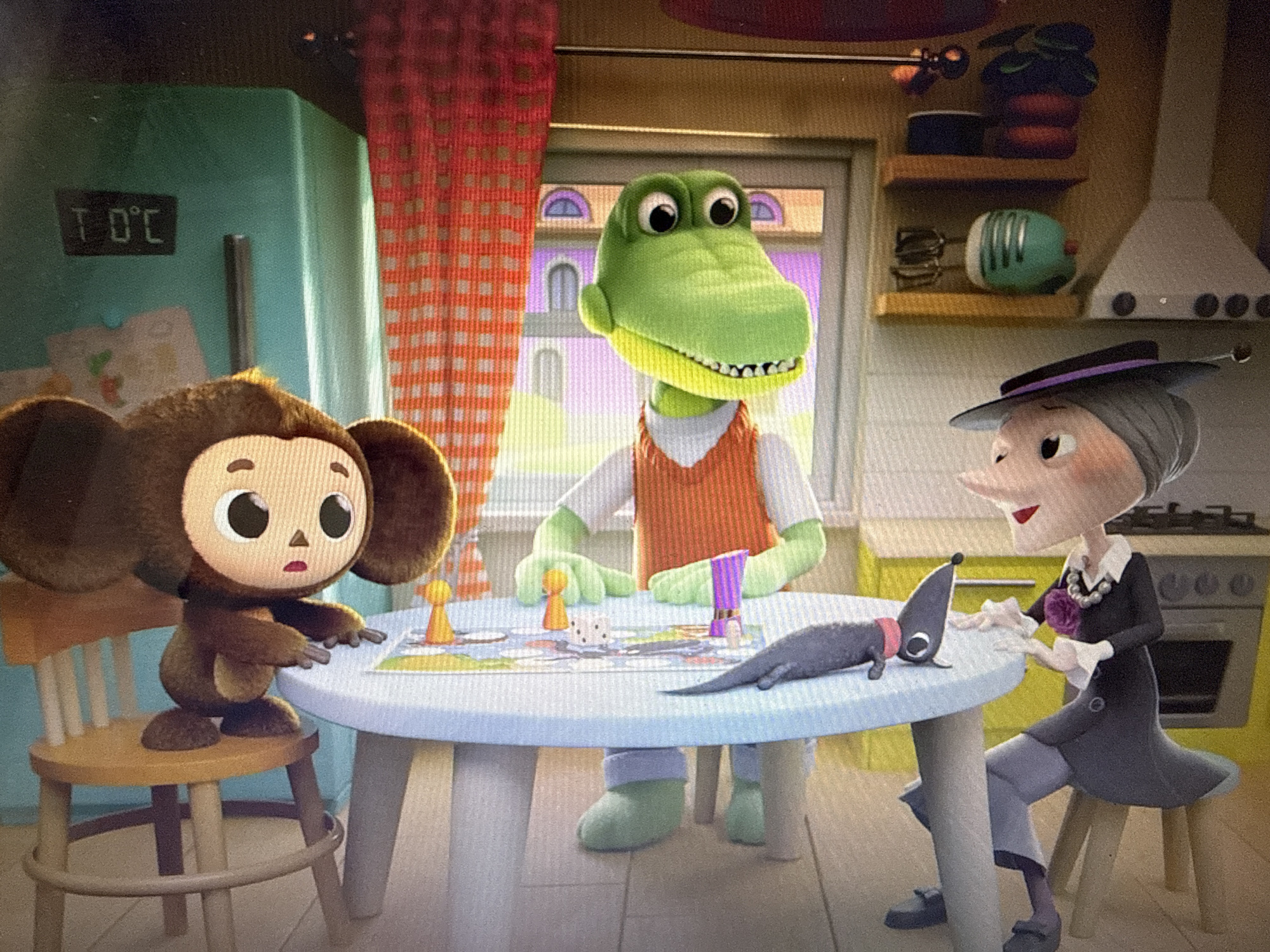 This image depicts an indoor scene, likely a kitchen, with three animated characters gathered around a round, light blue table. The characters include a fuzzy brown creature with large ears and expressive eyes, a green crocodile wearing an orange vest over a white shirt, and a woman with gray hair styled in a bun, wearing a black outfit, a purple hat, and a necklace with a flower-like pendant. A small, dark gray, dog-like creature with a red collar is also present on the table.
The table is set with a board game, including pawns, dice, and a purple marker. The characters appear engaged in playing the game. The background shows a refrigerator with a digital display showing "T 0 C," indicating 0 degrees Celsius. There is also a red and white checkered curtain, shelves with various kitchen items like a mixer and a plant, and a stove. The lighting suggests it is daytime. The scene evokes a sense of playful interaction and domesticity. The setting appears to be a stylized, possibly cartoonish, representation of a kitchen in Pushkino, Russia.