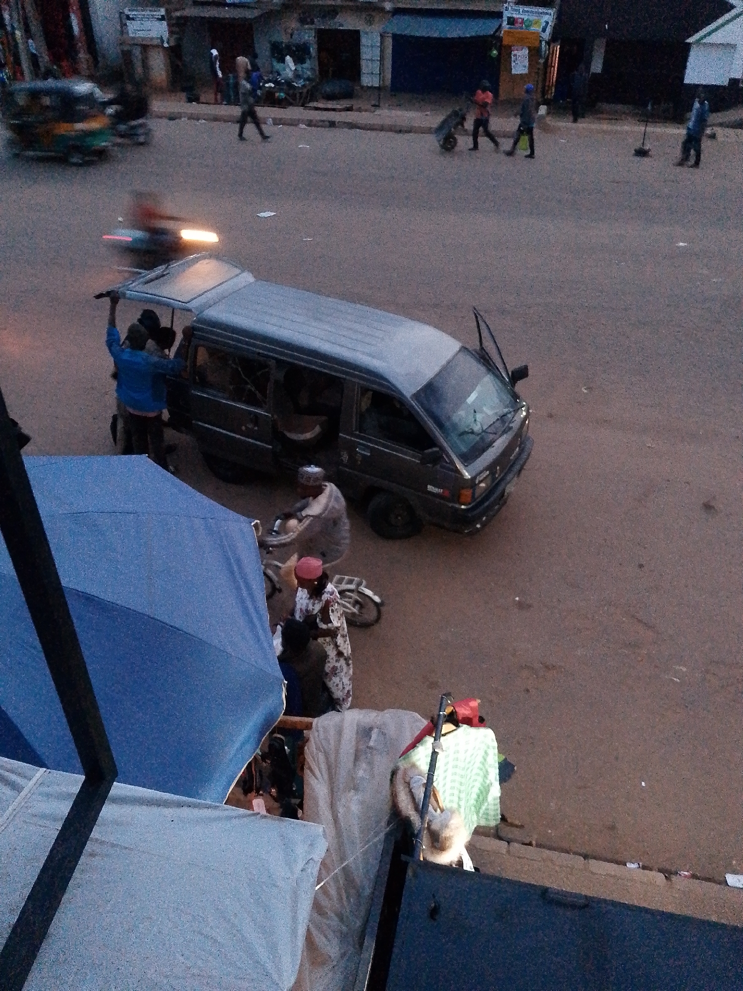 The image depicts a street scene in Bukuru, Nigeria, during dusk or early morning. A dark-colored van with its rear doors open is parked on the asphalt road. A person in a blue shirt is standing near the rear of the van, with their arm raised. Two other individuals are visible inside the van's cabin.
In the foreground, a large blue umbrella is partially covering a stack of white fabric. To the right of the umbrella, a woman wearing a patterned dress and a red headscarf is standing near a bicycle. Another person, possibly a man, wearing a cap and traditional attire, is seated on a bicycle to the left of the woman. Further in the background, a few more people are walking on the side of the road, and a motorcycle is partially visible. The overall environment suggests a bustling marketplace or residential area.