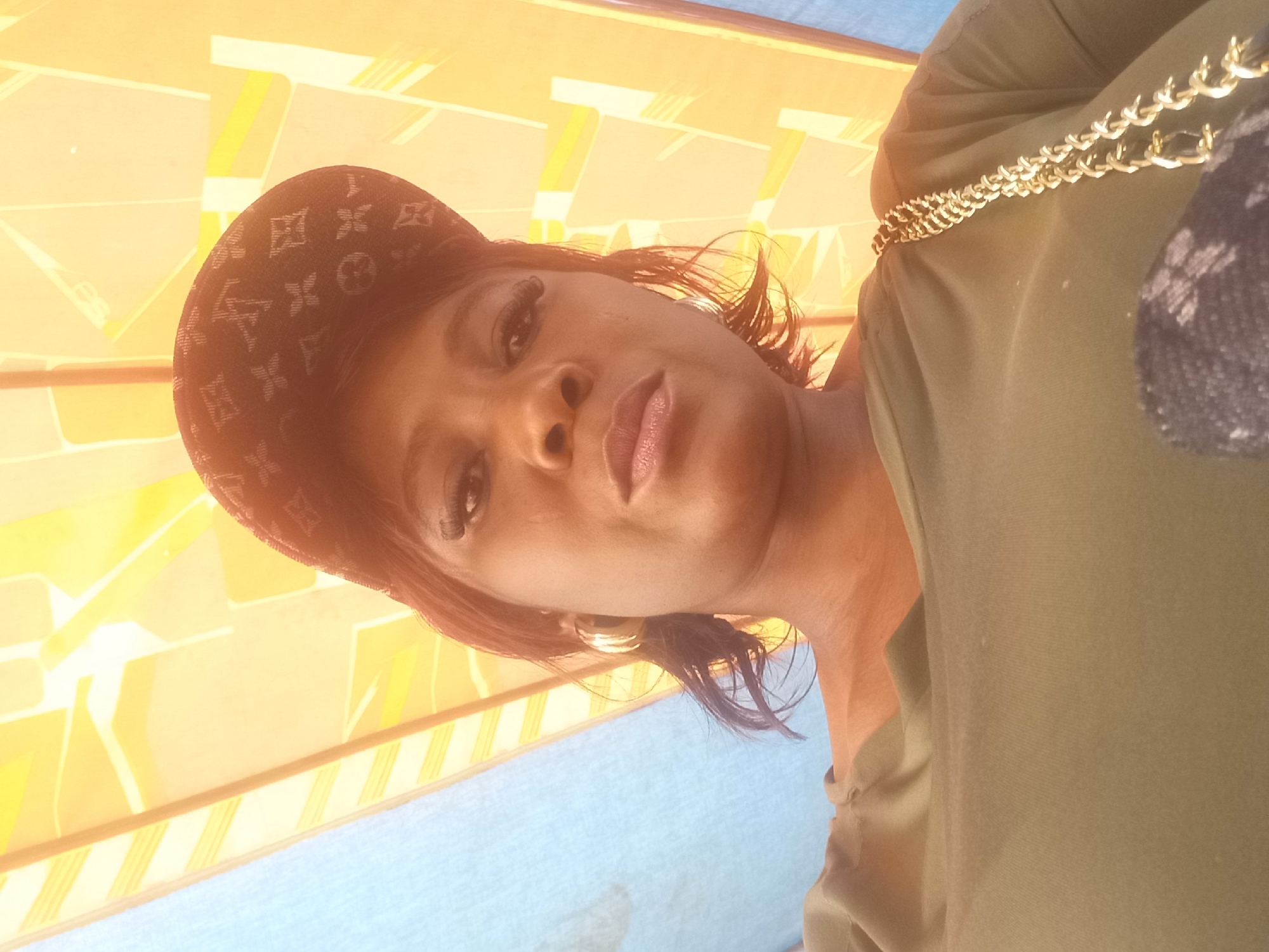A woman wearing a green off-the-shoulder shirt and a patterned baseball cap poses for a selfie outdoors. She has gold hoop earrings and long false eyelashes. A gold chain purse is visible on her right shoulder. The background consists of a blue tarp and an orange and yellow awning. The lighting suggests it is daytime. The specific location in Mangu Town, Nigeria cannot be determined from the image.