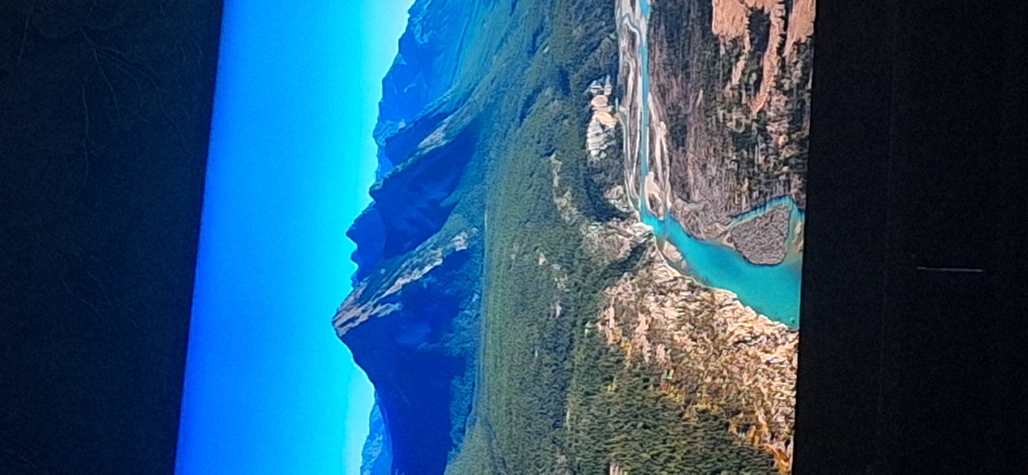 The image is a photograph of a vast mountain landscape under a clear blue sky. The foreground features a vibrant turquoise river winding through a rugged canyon with rocky, barren slopes. Dense green forests cover the lower parts of the mountains, transitioning into darker, more shadowed slopes. In the background, towering, rugged mountains with steep, rocky faces dominate the horizon, their peaks catching the sunlight. The overall impression is one of natural grandeur and untouched wilderness. There is no discernible connection to the city of Bila Tserkva, Ukraine, or any urban environment.