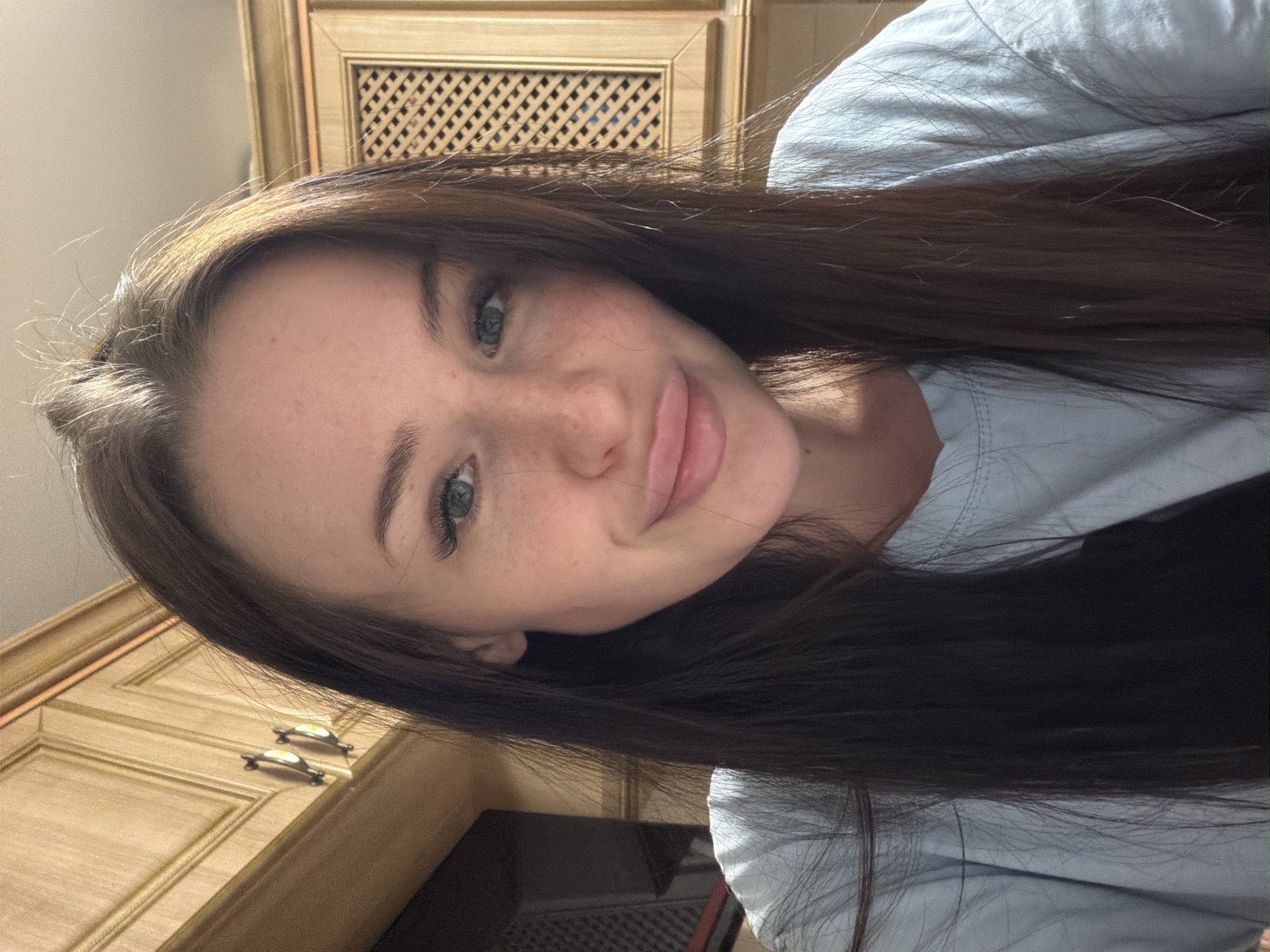 A close-up portrait shows a young woman with long, dark brown hair that cascades over her shoulders. She has fair skin, light-colored eyes (appearing blue or green), and a scattering of freckles across her nose and cheeks. Her lips are slightly parted in a soft, subtle smile. She is looking directly at the camera, and her head is tilted to the side, giving the impression of a selfie or casual portrait. She is wearing a light blue or light grey long-sleeved shirt.
The background consists of light-colored wooden cabinetry or furniture, suggesting an indoor setting like a kitchen or a room with built-in storage. One section of the furniture features a decorative, gridded or slatted panel. Another part of the wooden structure visible towards the bottom left has two small, metallic drawer pulls or handles. The lighting appears to be artificial or diffused, illuminating her face and hair. No visible text is present in the image.