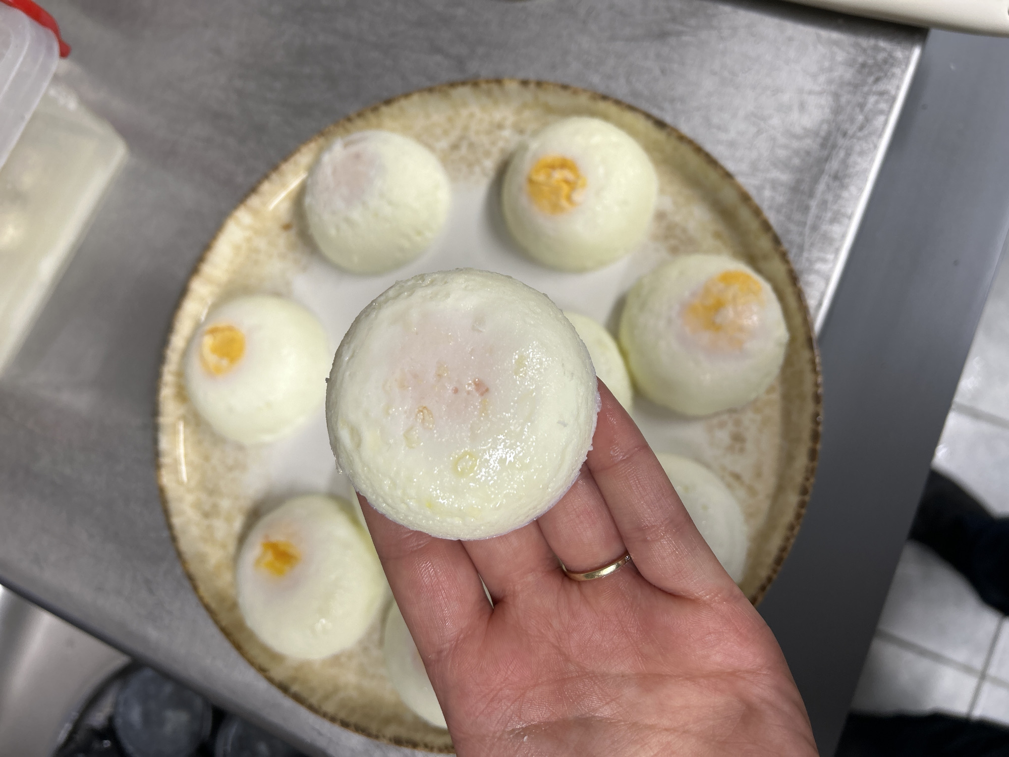 A high-angle shot shows a hand holding a white, round, poached egg. The egg has a light orange yolk visible through a thin layer of white. The hand has a gold ring on its ring finger. On the patterned plate beneath, there are seven more of the same poached eggs. The background consists of a stainless-steel surface, a white container with a red lid, and a hint of black clothing. No information suggests this is in Burgas, Bulgaria.