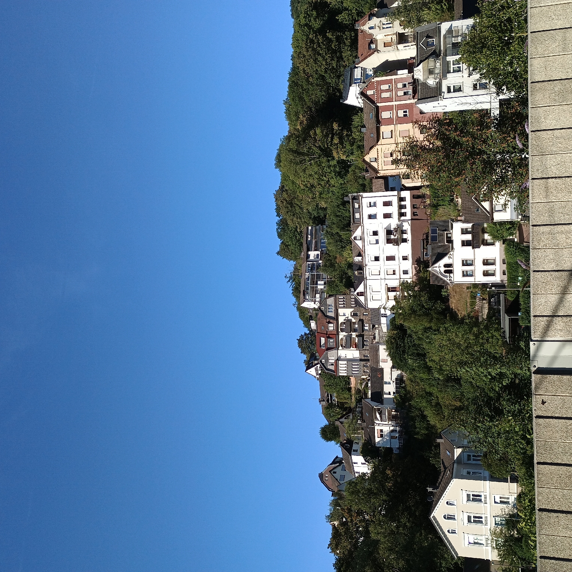 The image shows a view of houses built on a steep hillside in Altena, Germany. The sky is clear and blue, indicating a sunny day. The houses are of varying architectural styles and colors, with some featuring red tile roofs and others gray or dark roofing. Many of the houses have balconies and windows with shutters. The hillside is densely covered with trees, suggesting a natural or park-like setting surrounding the residential area. In the foreground, a concrete wall or barrier runs horizontally across the bottom of the frame, partially obscuring the view of the lower houses and the ground. There are no people or vehicles visible in the image. The overall scene depicts a residential neighborhood situated on a challenging, sloped terrain.