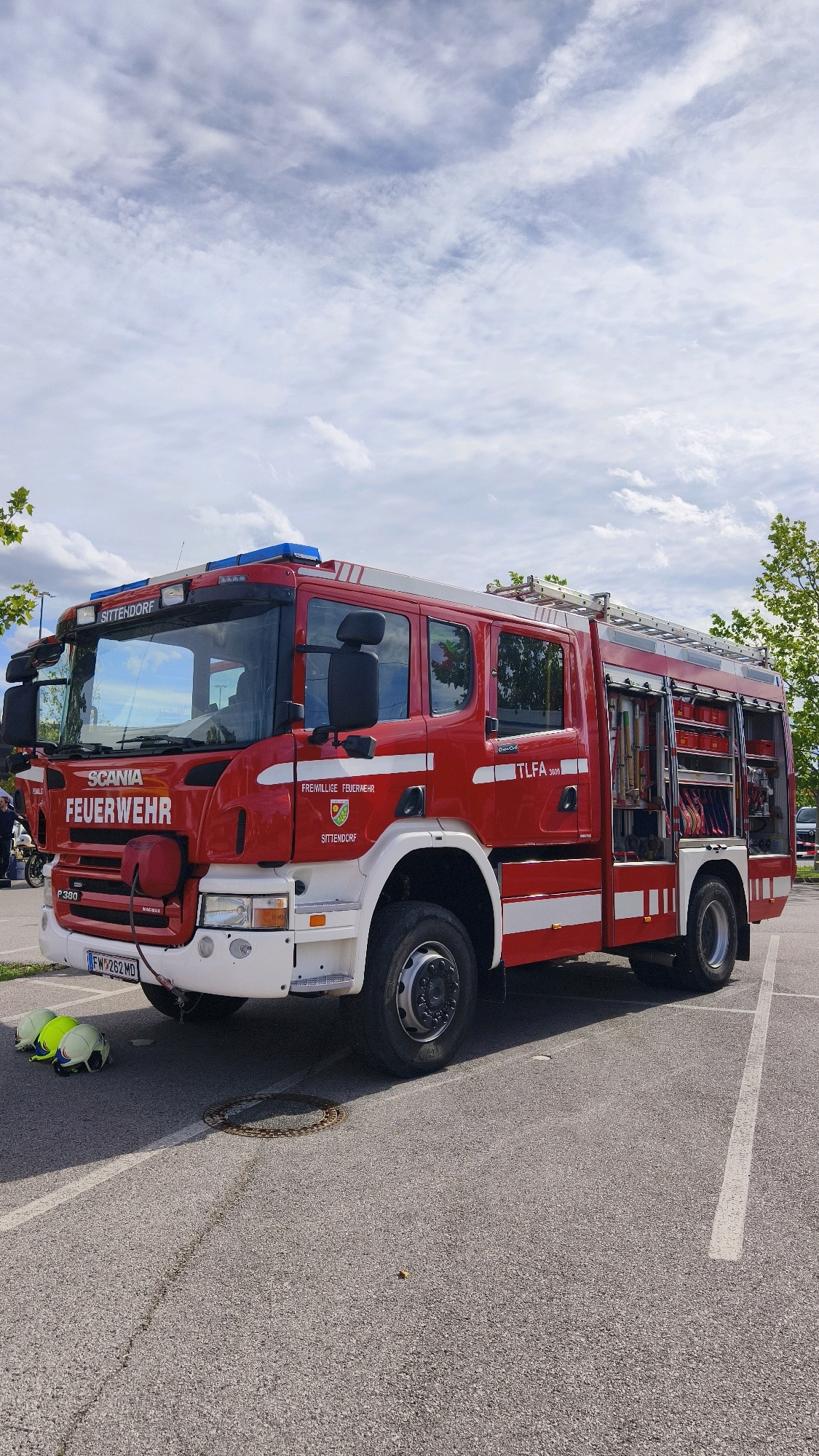 A red and white Scania Feuerwehehr fire truck is parked on an asphalt surface, likely a parking lot, under a cloudy sky. The fire truck, identified as a TLFA 3000 and belonging to the Freiwillige Feuerwehr Sittendorf, is oriented towards the viewer with its front left side visible. The truck features a ladder on its roof and compartments on its right side filled with equipment. In the foreground, three fire helmets are placed on the ground near the front left wheel of the truck. A manhole cover is visible on the asphalt in front of the truck. The background includes trees and some parked vehicles. The scene appears to be an outdoor event or demonstration in or near Sittendorf, a municipality within the district of Mödling, Lower Austria, which is part of the greater Vienna metropolitan area, often associated with Vösendorf due to its proximity.