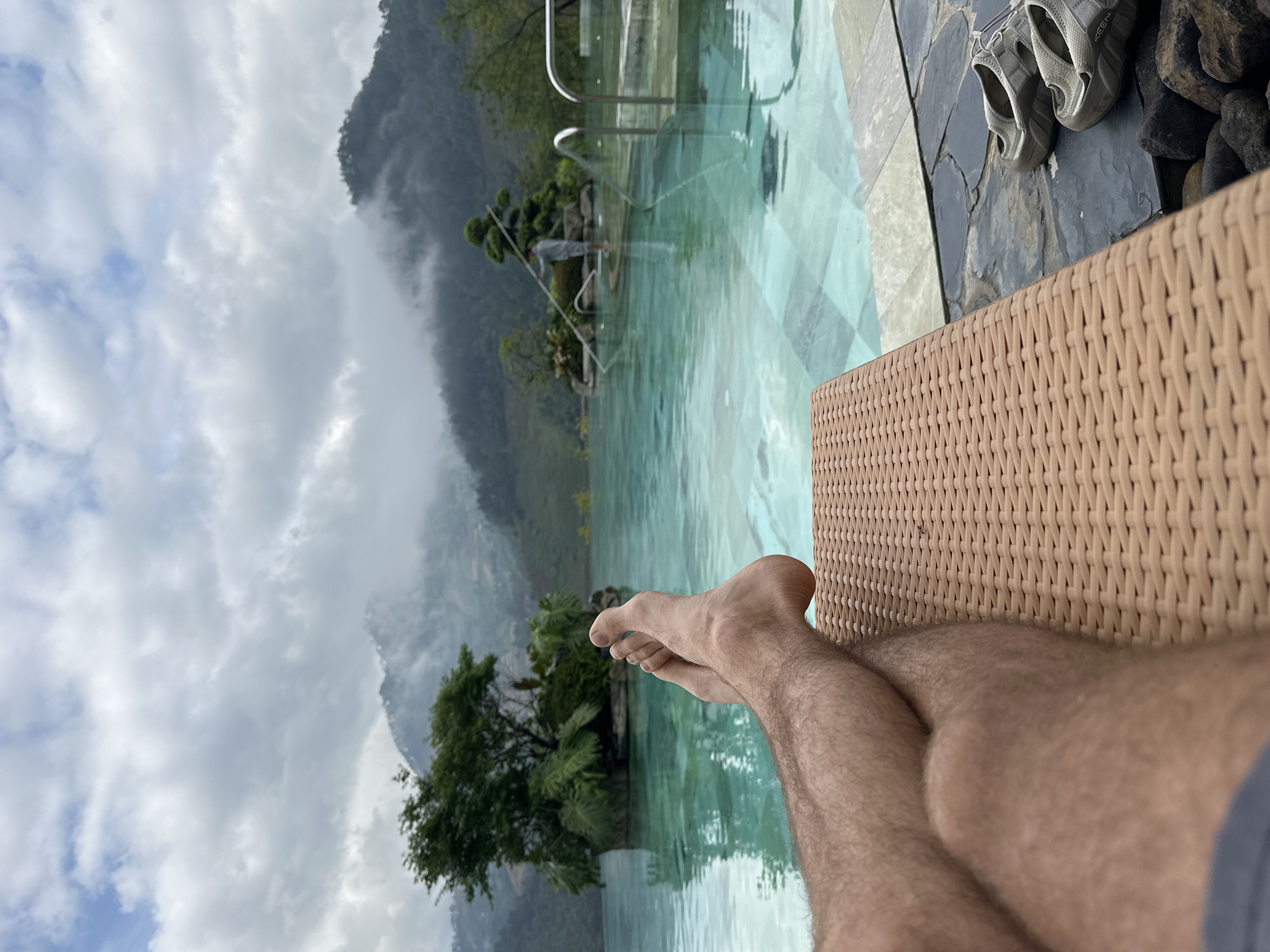 An overhead perspective captures a person's legs and feet resting on a lounge chair, overlooking an infinity pool and a mountainous landscape in Púng Luông, Vietnam.
In the immediate foreground, a small section of dark grey fabric, likely shorts, is visible at the bottom left. Above this, a pair of bare, hairy legs are crossed at the ankles, resting on a light brown, woven-textured chaise lounge chair that occupies the lower right quadrant of the image. The feet are positioned towards the left. To the right of the lounge chair, on a dark, irregular stone surface, a pair of light grey "Keen" brand sandals are placed. Below the sandals, several dark, smooth river rocks are visible.
The midground features a turquoise-blue infinity pool with visible patterned tiling on its floor. The pool water reflects the surrounding environment. Within the pool, on the left side, a natural-looking island densely covered with green trees and ferns is present. Towards the right side of the pool, another smaller, manicured island features a singular, sculptural tree. A person, dressed in light-colored attire, stands in the pool between the right island and the pool edge, holding a long, light-colored pole, possibly for maintenance. Chrome-colored handrails are visible at the pool's edge on the far right.
The background consists of rolling green mountains, partially enveloped in mist and low-hanging grey and white clouds. Patches of lighter blue sky are discernible through the cloud cover. The overall atmospheric condition appears overcast.