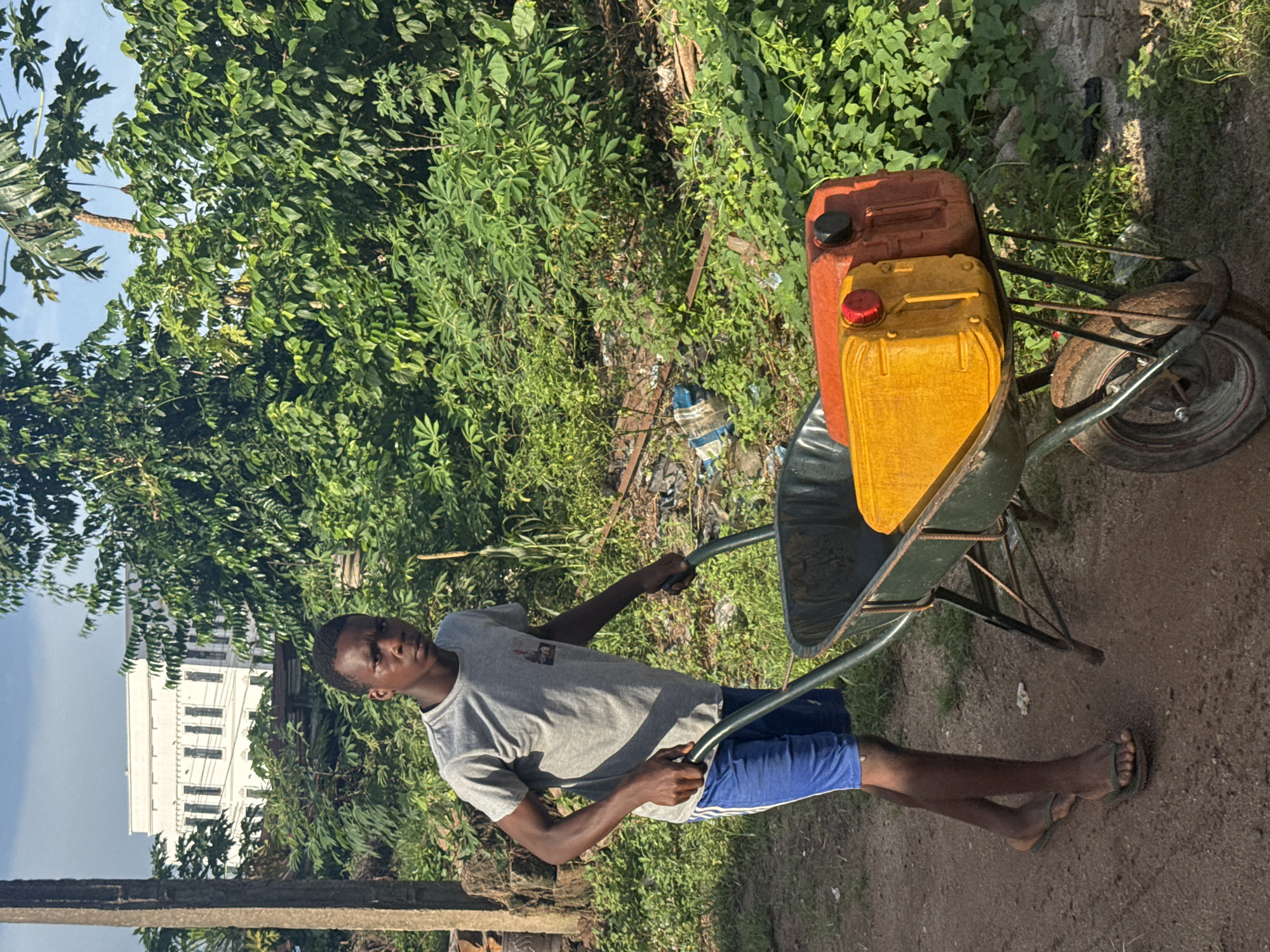 A medium, eye-level shot features a young African man pulling a wheelbarrow with two plastic jerrycans in Aboh, Nigeria. The man, dressed in a grey t-shirt and blue shorts, is pulling the dark-green wheelbarrow along a dirt path. The wheelbarrow contains a red and a yellow jerrycan. The wheelbarrow’s tire shows wear. Lush, green vegetation fills the background. A white building is visible behind the vegetation. The sky is cloudy.