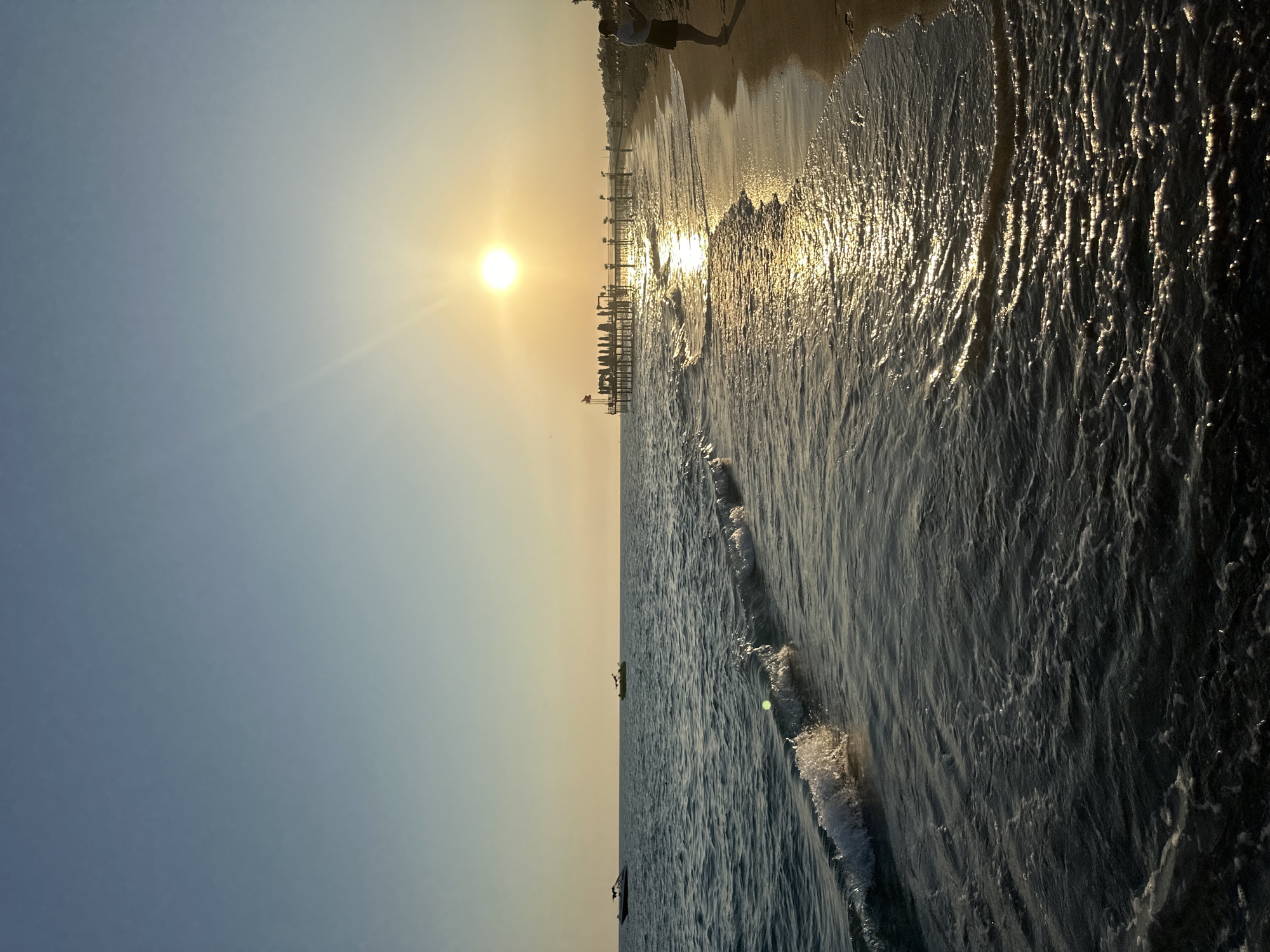 This image captures a serene sunset scene at a beach in Manavgat, Turkey. The sun, a bright orb of golden light, is low in the sky, casting a warm glow across the tranquil sea. Its reflection shimmers on the water's surface, creating a path of light towards the shore. Gentle waves lap at the sandy beach, leaving a foamy trail as they recede.
In the distance, a wooden pier stretches out into the water, lined with what appear to be lounge chairs and umbrellas, suggesting a resort or public beach area. Two boats are visible on the sea, one closer to the shore and another further out. A single person stands on the beach, facing the water, seemingly lost in contemplation of the beautiful view. The sky transitions from a pale blue at the top to a soft yellow-orange near the horizon, a typical palette for a sunset. The overall atmosphere is one of peace and natural beauty.