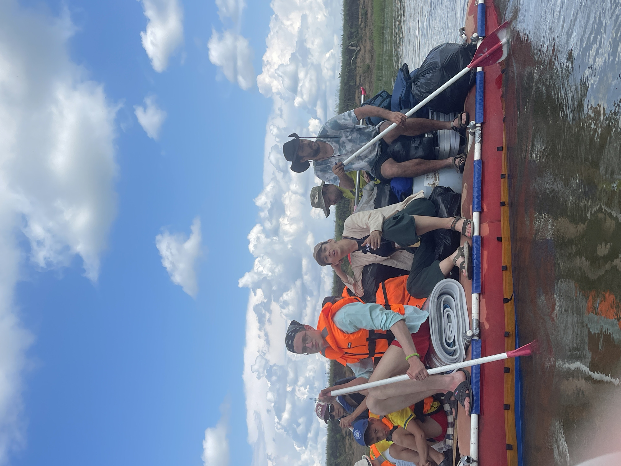 A group of people are on a raft on a body of water under a blue sky with white clouds. The raft is red with yellow and blue accents. Several people are wearing orange life jackets. One person in the foreground on the left is wearing a blue shirt, red shorts, and an orange life jacket, and is holding a paddle. Another person in the foreground on the right is wearing a camouflage shirt and a black cowboy hat, and is also holding a paddle. There are several bags and rolled-up items on the raft. The background shows a grassy bank with some vegetation. The scene appears to be a recreational activity on a river or lake.