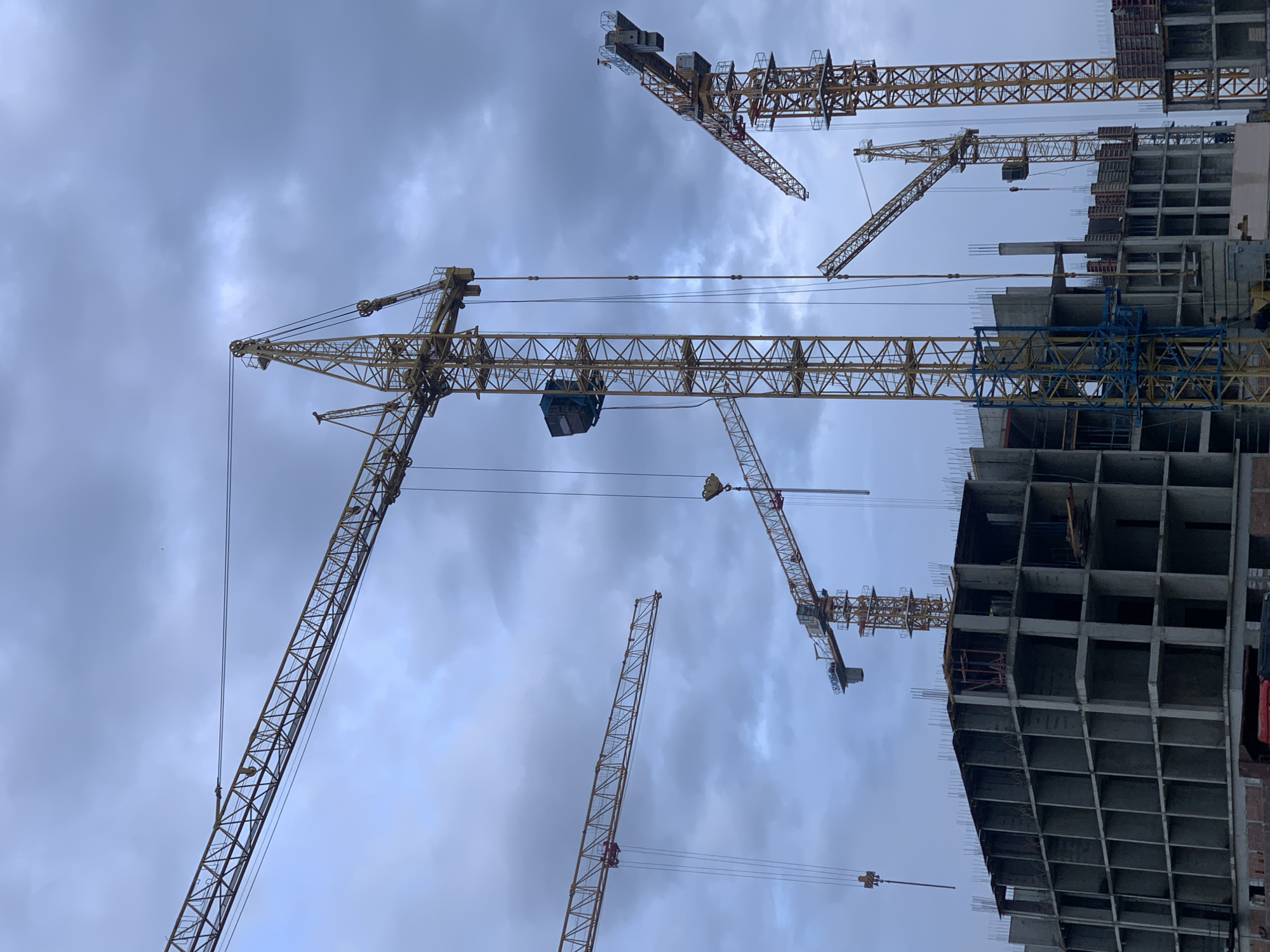 The image captures a construction site with multiple tall yellow tower cranes against a cloudy, overcast sky. The cranes are the primary objects, with their intricate lattice structures dominating the frame. One crane is in the foreground, its long arm extending diagonally upwards. Another crane is seen in the middle ground, positioned near a partially constructed building. In the background, more cranes are visible, some partially obscured by the building.
The building itself is a multi-story concrete structure, showing exposed rebar and concrete floors. It appears to be in an early to mid-stage of construction. The cloudy sky suggests it is either morning or late afternoon, or possibly a generally overcast day. There are no people visible in the image. The scene conveys a sense of ongoing development and construction. The location is identified as Stavropol, Russia. There is no visible text in the image.