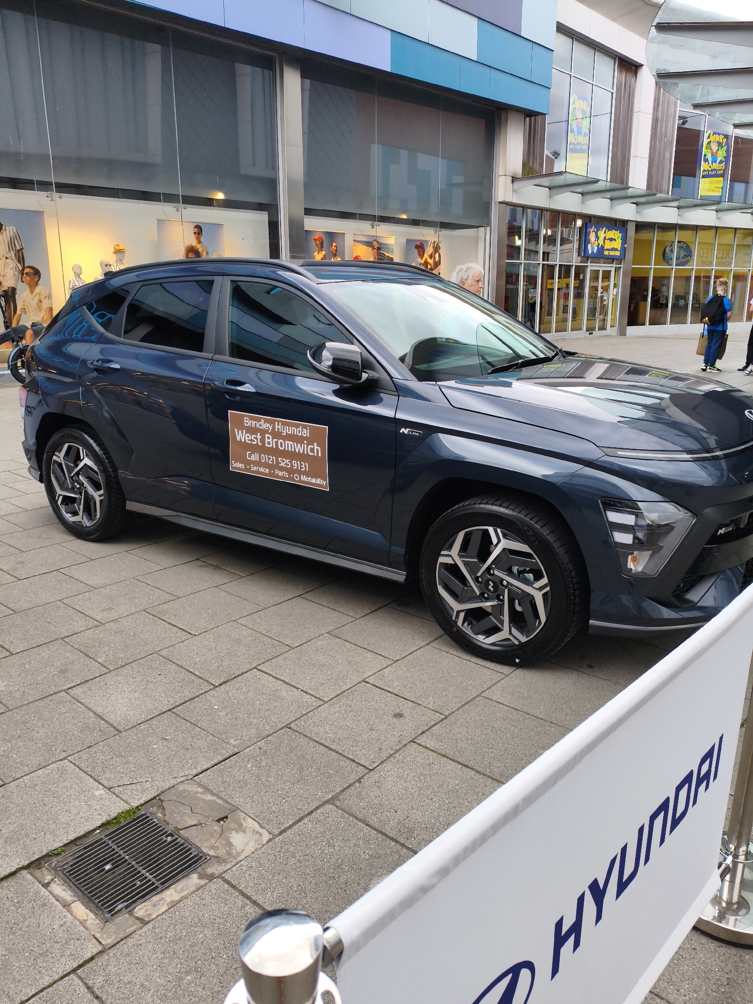 A dark blue Hyundai Kona N Line is parked on a paved pedestrian area in front of a row of shopfronts. A large white banner with a blue Hyundai logo and text is partially visible in the foreground. The car has a sticker on its driver's side door that reads "Brindley Hyundai West Bromwich" and provides a contact phone number and service details. Behind the car, shop windows display mannequins and merchandise. In the background, other shops with signage, including "Monkey Monkeys" play centre, are visible. Several people are walking in the background, including a person with a blue backpack. The environment appears to be a public walkway or town centre, likely in Sandwell, United Kingdom, given the mention of West Bromwich on the car's sticker.
