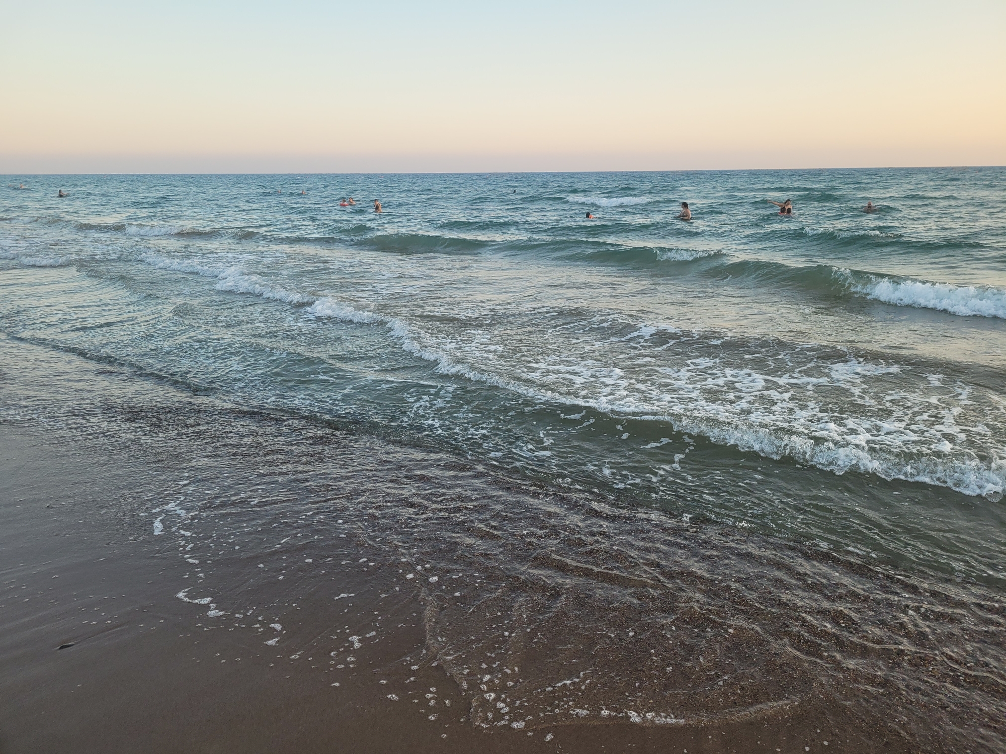 The image depicts a serene beach scene with a calm sea under a pale sky. Several people are visible in the water, swimming or wading. The foreground shows the wet sand of the beach, with gentle waves lapping at the shore, creating small patterns of foam and ripples on the sand. The sea itself is a tranquil blue-green, with small waves breaking and creating whitecaps further out. The horizon is a clear line where the sea meets the sky, which is a soft gradient of pale blue and pink, suggesting either early morning or late evening. The scene is devoid of any specific landmarks that would pinpoint it to Episkopi Lemesou in the United Kingdom, as Episkopi Lemesou is located in Cyprus, not the UK.