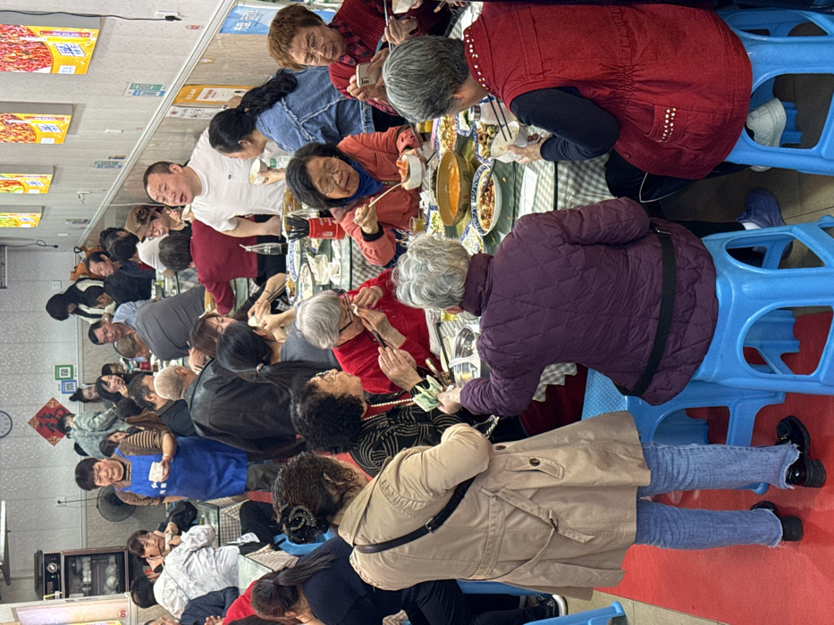This image depicts a bustling indoor restaurant in Yuzhong District, China. The establishment features white walls, overhead fluorescent lighting, and a red floor runner in the foreground. Multiple round tables, each covered with a light blue and white checkered tablecloth, are occupied by groups of diners. Seating consists primarily of blue plastic stools.
Approximately twenty to thirty individuals are visible, spanning various age groups but with a notable presence of older adults. At the forefront, a woman in a light tan trench coat, blue jeans, and black loafers stands with her back to the camera, holding a bundle of green banknotes in her right hand, extending them towards another woman seated at a table. This seated woman, with short grey hair and a purple quilted jacket, reaches her left arm out to receive the cash. Beside her, another woman in a red vest with white decorative studs is seated, holding a white bowl with chopsticks.
The tables are laden with numerous dishes, including bowls of soup, various prepared foods, small white rice bowls, and sets of chopsticks. People are actively engaged in eating, conversing, and socializing. One older woman in a red top and glasses is shown looking at a smartphone while eating. Another woman in an orange jacket is using chopsticks to eat from a bowl of red-colored food.
In the mid-ground, a server, a middle-aged woman wearing a blue apron over a striped shirt, walks between tables holding a white bowl. Further back, a man in a white t-shirt is visibly speaking, his mouth open, while holding a small white bowl. Other diners occupy various tables, some looking towards the camera, others engaged in their meals or conversations. The background shows more patrons, partially obscured, and the restaurant walls decorated with framed pictures, posters featuring Chinese characters and vibrant food imagery, and a wall-mounted clock.