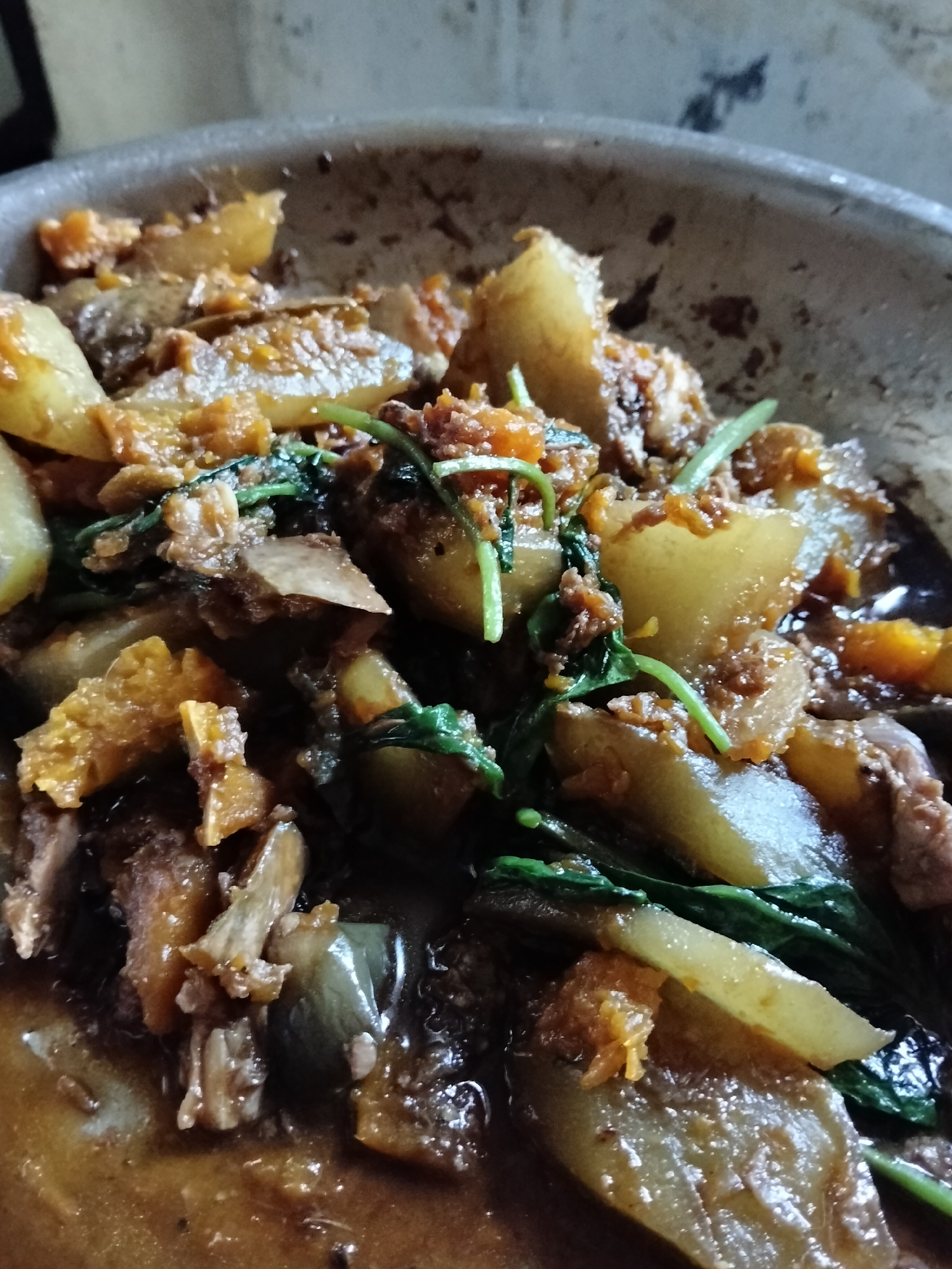 The image shows a close-up view of a pan filled with a cooked dish. The dish appears to be a stew or braise, with visible chunks of meat and vegetables in a rich, dark brown sauce. Some of the vegetables appear to be eggplant and possibly squash or potato, cut into irregular pieces. Green leafy vegetables are also interspersed throughout the dish. The food is presented in a metallic pan, likely a wok or a similar cooking vessel. The lighting suggests an indoor setting, possibly a home kitchen. There is no indication of the specific location within Davao City, Philippines, beyond the general knowledge that this is a food item likely prepared and consumed there. The image focuses solely on the food and the pan.