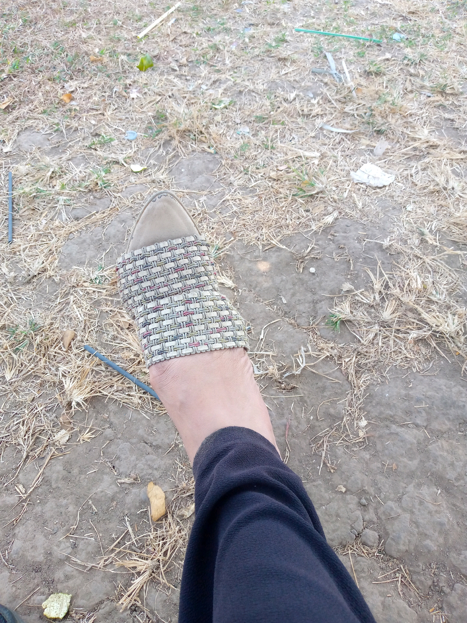 A first-person perspective shot shows a person's left foot, partially covered by a black garment, resting on dry, dusty ground scattered with straw and a few green shoots of grass. The foot is adorned with a light brown sandal featuring a woven strap with a pattern of light brown, white, and red threads. Two thin, dark gray rods lie on the ground near the foot, one running parallel to the sandal strap, and another angled across the foreground. In the background, more dry straw and dirt are visible, along with scattered debris, including a small piece of green material, a crumpled white object, and some dried leaves. The scene appears to be an outdoor environment, possibly a park or an uncultivated area within the city of Pacing, Indonesia.