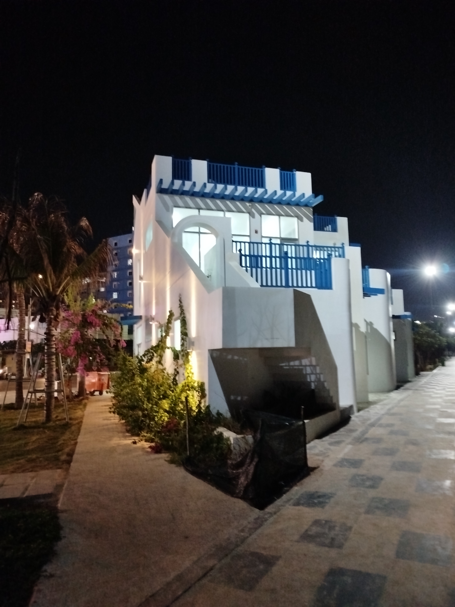 Captured at night in Ho Chi Minh City, Vietnam, the scene features a multi-level white building with blue accents. The building, possibly residential or commercial, exhibits a modern architectural style. To the left, palm trees and flowering plants line a pathway, while on the right, a tiled walkway extends into the distance, illuminated by streetlights. A dark covering obscures a portion of the area near the building's base. Additional structures are visible in the background. No people are visible.