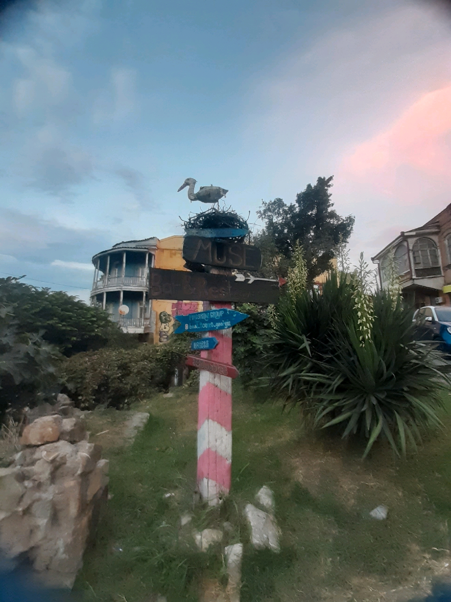 A weathered wooden signpost, painted with alternating pink and white diagonal stripes, stands in the foreground. Atop the signpost is a nest with a decorative bird. Several directional signs are attached to the post; one reads "MUSEUM" in dark lettering on a wooden plank, pointing left. Below this, a blue arrow points left, with faint text that appears to be in Georgian script. Another blue arrow points right, also with Georgian script. A pink sign with white text is visible at the bottom.
Behind the signpost, a building with a distinctive curved, two-story balcony painted in light blue stands out. The building has a yellow facade. Further to the right, another building with arched windows and darker trim is visible, with a car partially obscuring its lower section.
The environment is grassy with some scattered rocks and a large yucca plant with white flowers to the right of the signpost. The sky above is a mix of pale blue and soft pinkish hues, suggesting it might be dusk or dawn. The scene appears to be in an outdoor setting, possibly a street or garden area in Tbilisi, Georgia, given the architectural style and the visible Georgian script.