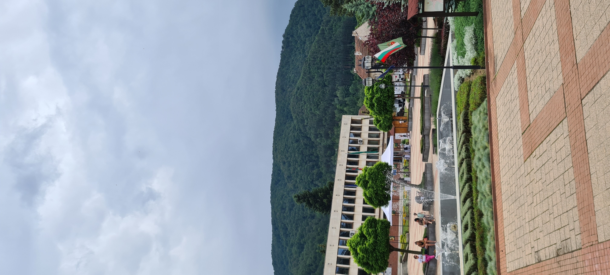 The image depicts the central plaza of Etropole, Bulgaria, under an overcast sky. A prominent beige, five-story building with multiple dark-framed windows occupies the left-center, displaying five flags on its facade, including at least one Bulgarian flag. Behind this building and extending across the upper background, a densely forested mountain rises under the grey, cloudy sky.
In the mid-ground, a paved public square features a rectangular multi-jet fountain system. Surrounding the fountain are meticulously manicured green bushes, some of which are shaped to spell out the letters "ETROPOLE" in Cyrillic. Several individuals are present in the square: four people are seated on a bench to the left of the fountain, and a small group, including a child, is visible near the fountain's center. To the right of the fountain, a black lamppost displays a Bulgarian flag and a green flag with an emblem. Two white event tents are erected in the plaza behind the fountain, and several small, topiary-style trees are planted throughout the landscaped areas. The foreground consists of light-colored paving stones with darker red-brown stone accents forming a grid pattern.