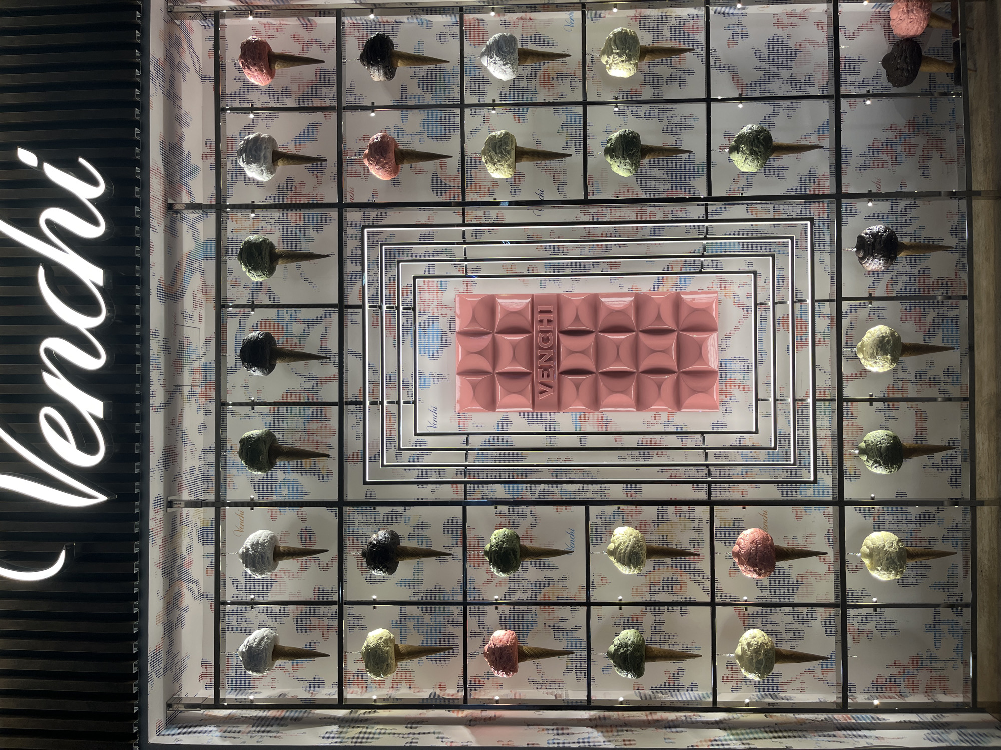 A retail storefront display for "Venchi" is depicted in Bologna, Italy. The upper section of the display features the brand name "Venchi" in illuminated white script against a dark, vertically grooved background. The main display area consists of a grid-like structure with dark, slender vertical and horizontal bars.
Within this grid, numerous decorative ice cream cones are suspended individually by thin wires. These cones feature light brown wafer-cone bases and sculpted "scoops" in various colors: off-white, dark chocolate brown, light mint green, pale yellow, and strawberry pink. There are 35 such cones visibly arranged in 7 vertical columns and 5 horizontal rows, with varying colors distributed across the grid. The background behind the cones is a light-colored wall adorned with a patterned graphic featuring small, irregular shapes in red, blue, black, and white.
To the right of the display's center, a large, rectangular, solid pink object is prominently featured. This object, textured with a grid of raised squares, resembles an oversized chocolate bar. The word "VENCHI" is embossed horizontally across its middle in a slightly lighter pink or white. This pink slab is framed by a series of concentric rectangular frames made of thin, illuminated white lines, creating a glowing border around the central display element. The overall lighting is bright, illuminating the display elements and the brand signage. No individuals are present in the scene.