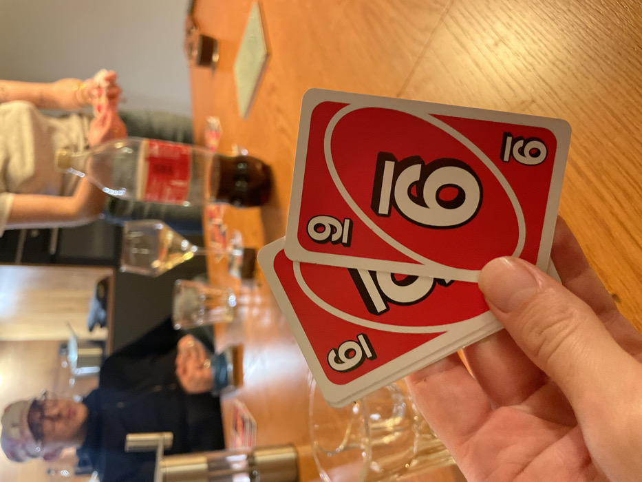 A person holds two red UNO cards with the number six in their hand. A wooden table is in the background with multiple people playing the game. There are also glasses and a Coke bottle on the table. One person wearing glasses is in the lower left corner of the shot.