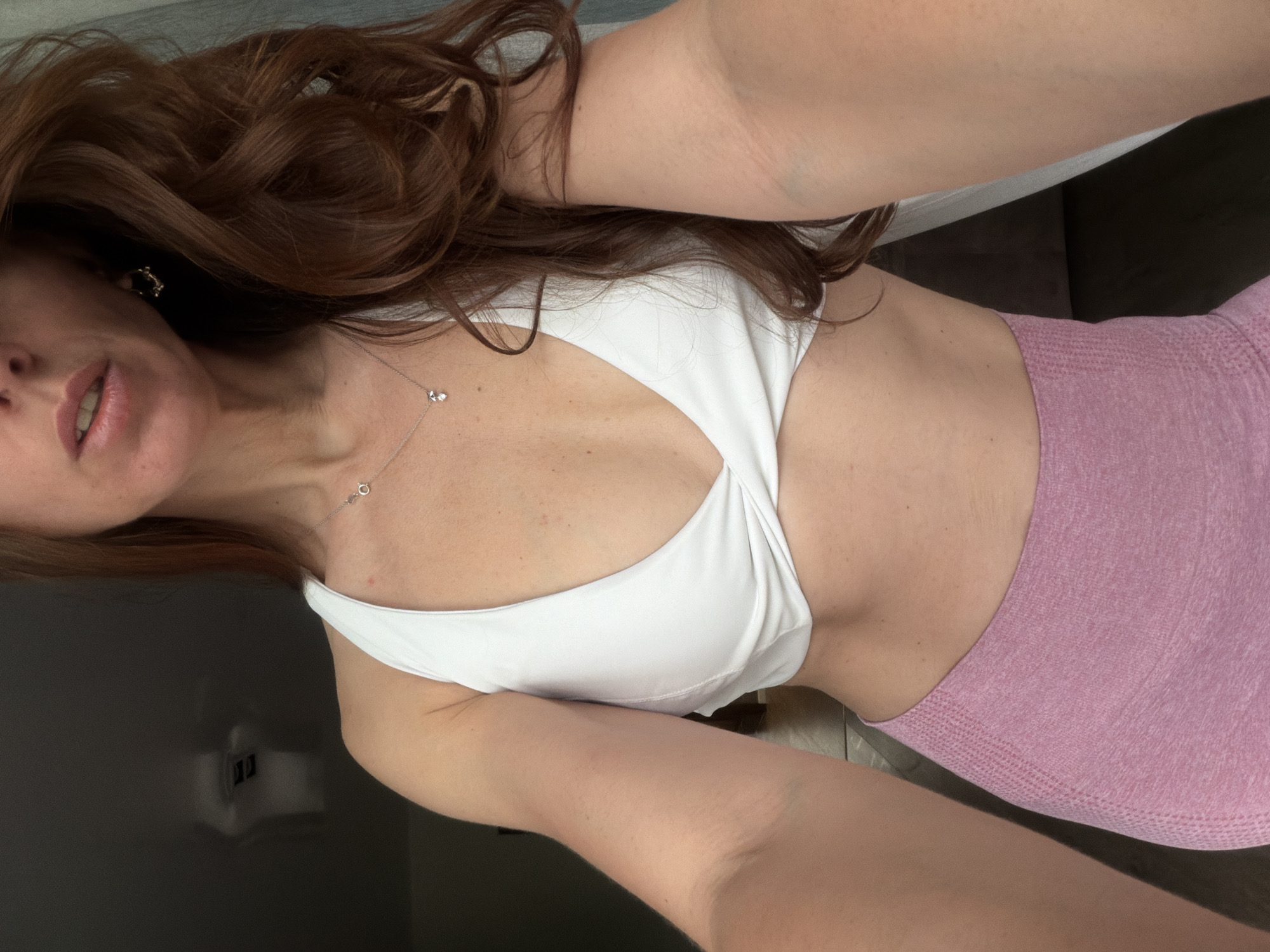 This is an indoor close-up selfie of a person, likely a woman, positioned from the chest up with her head rotated to the left. She has long, voluminous reddish-brown hair that frames her face and shoulders. A portion of her face is visible on the left, including her chin, mouth slightly agape to reveal teeth, and part of her left cheek and eye. She wears a small, dark earring on her left ear.
She is dressed in athletic wear, consisting of a white sports bra or cropped top with a twisted knot design at the front, and textured, high-waisted pink leggings or shorts. A delicate silver chain necklace with two small pendants rests on her chest, which shows some freckles. Her skin tone is fair. Her left arm extends across the top of the frame, presumably holding the camera for the selfie, while her right arm is also visible.
The background is generally dark and indistinct, suggesting an indoor setting. On the right, a dark, possibly upholstered surface is visible, and on the lower left, there is a reflective dark surface, possibly a screen or mirror, showing a vague reflection of what might be an internal light source. The lighting is soft and even, indicating an indoor environment, but no specific time of day is discernible. The overall mood appears relaxed. There are no visible location cues within the image to explicitly confirm the location of Birobidzhan, Russia, and no visible text.