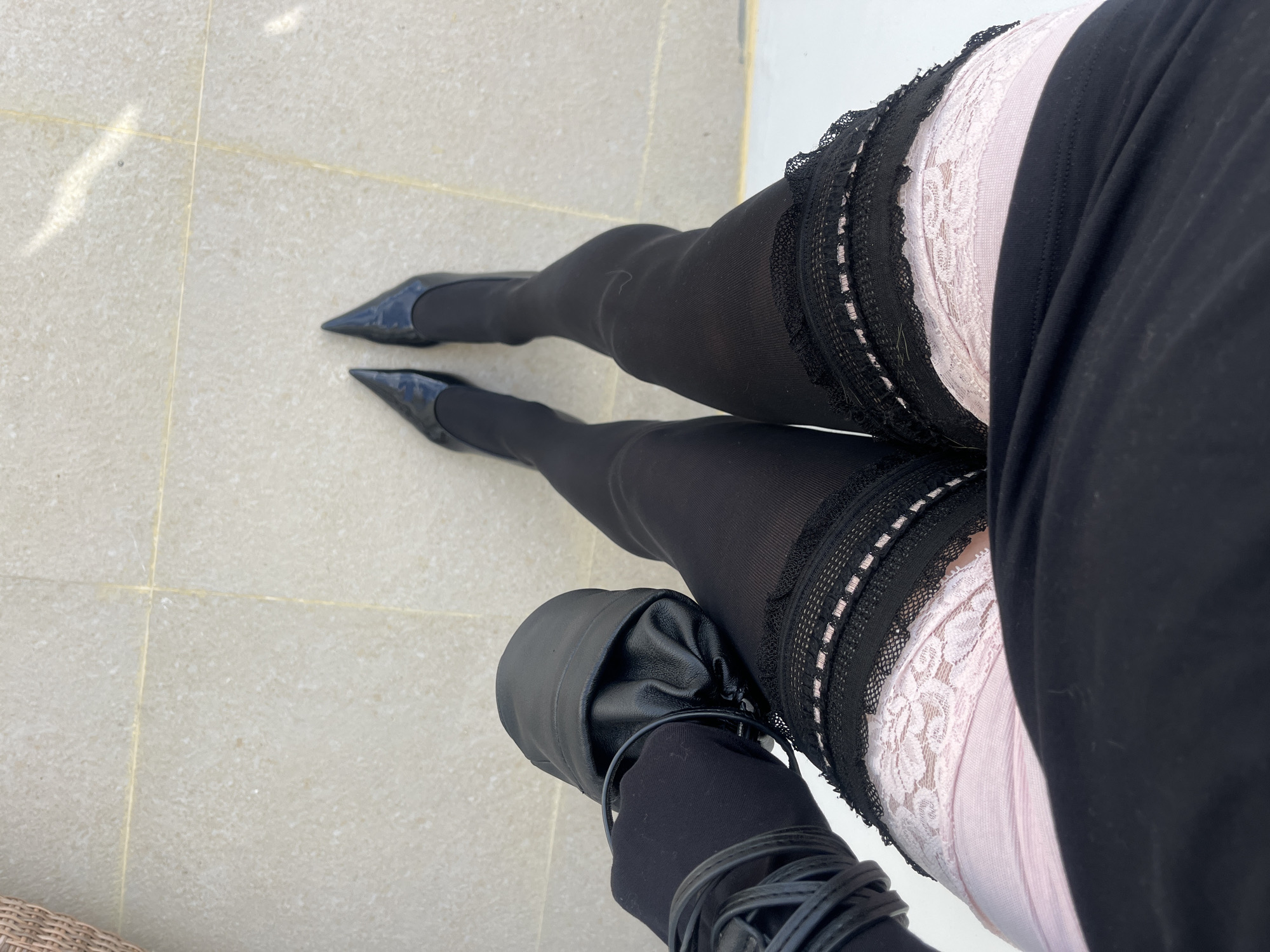 A high-angle, full body shot shows a person wearing black pantyhose, black heels, a pink and white lace-trimmed garter belt, and a black skirt. The person's right foot is placed inside a black leather bag with a drawstring. The person is standing on light beige tiles, possibly outdoors, in Kuta Selatan, Indonesia.
