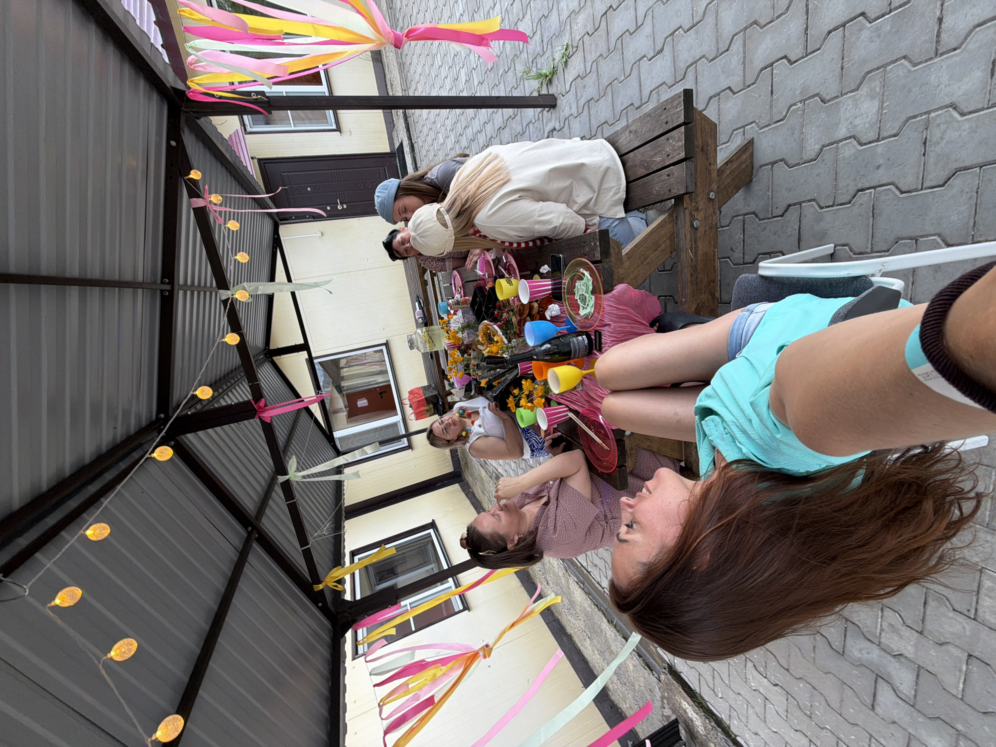 The image depicts a gathering of at least five women seated around a picnic table under a covered patio. The setting appears to be outdoors, with a tiled ground surface and a building visible in the background. Decorative streamers in various bright colors, including pink, yellow, and green, are suspended from the patio ceiling, along with string lights. The table is laden with food, drinks in colorful cups, plates, and bottles, suggesting a meal or celebration. The women are dressed casually, and their interactions suggest a social gathering. One woman in the foreground is in profile, looking upwards and smiling, with her legs extended. Another woman, seated at the table, is also looking in profile. The overall atmosphere seems relaxed and festive. The provided crops offer closer views of the string lights, streamers, and individuals at the table, aiding in detailed observation. The location is not explicitly stated within the image itself but the request specifies Birobidzhan, Russia.