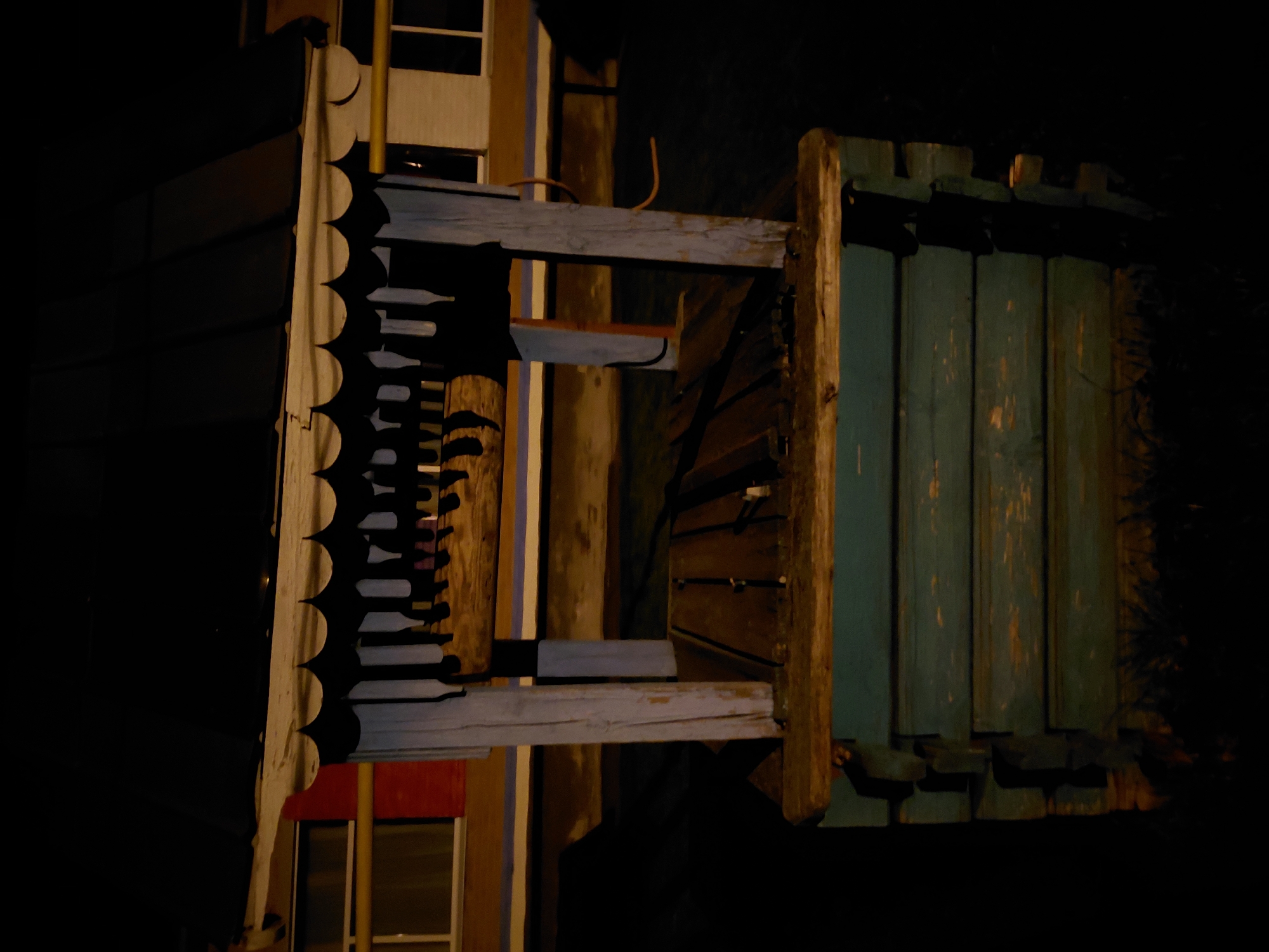 The image displays a close-up, dimly lit view of an outdoor structure, captured at night. The scene appears rotated; when viewed upright (rotated 90 degrees counter-clockwise), a building facade or large decorative element is presented.
On the left side, a section of a building features intricate, scalloped trim, often referred to as gingerbread detailing, likely made of wood and painted a dark color. Behind this trim, a lighter, possibly cream or beige, wall or panel is visible. Above, a black, indistinct architectural element sits, with a bright yellow pipe or conduit rising vertically near it. Below the scalloped trim, a flash of red is visible, perhaps an awning or a painted window frame.
Extending horizontally across the center of the frame is a weathered wooden beam, painted a light, possibly pale blue or grey, showing signs of wear and exposure. Above this beam, a curved orange wire or pipe is present.
To the right, a vertical wooden post supports a section composed of multiple wooden planks. These planks are painted a distressed, greenish-blue color, with chipped paint revealing lighter wood beneath, suggesting age and weathering. The bottom edge of this plank section features small, dark, rectangular elements, possibly decorative or structural.
The background is dark, indicating nighttime, with artificial light sources illuminating the structure and casting subtle glows on the visible elements. Hints of windows with frames or blinds can be seen behind the main structure. There is no discernible activity or visible text. The overall impression is of an aged, potentially rustic, or handcrafted architectural detail viewed in low light.