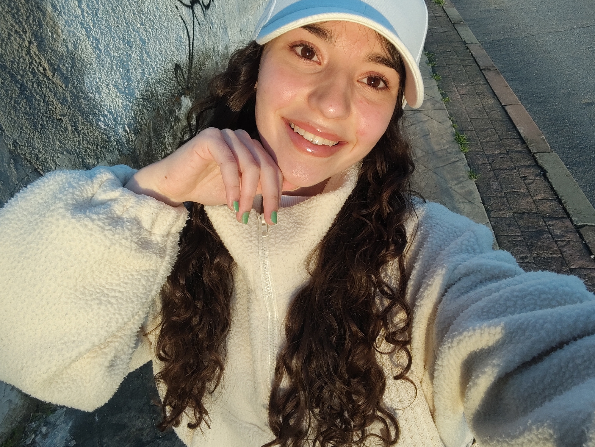 A young woman is taking a selfie outdoors. She is wearing a white baseball cap and a white fleece jacket with a zipper. Her hair is long, curly, and brown. She is smiling and has light-colored nails. The background includes a textured wall with graffiti, a street, and a sidewalk. The lighting suggests it's late afternoon or early morning. There is no explicit evidence that the image was taken in Sliven, Bulgaria.