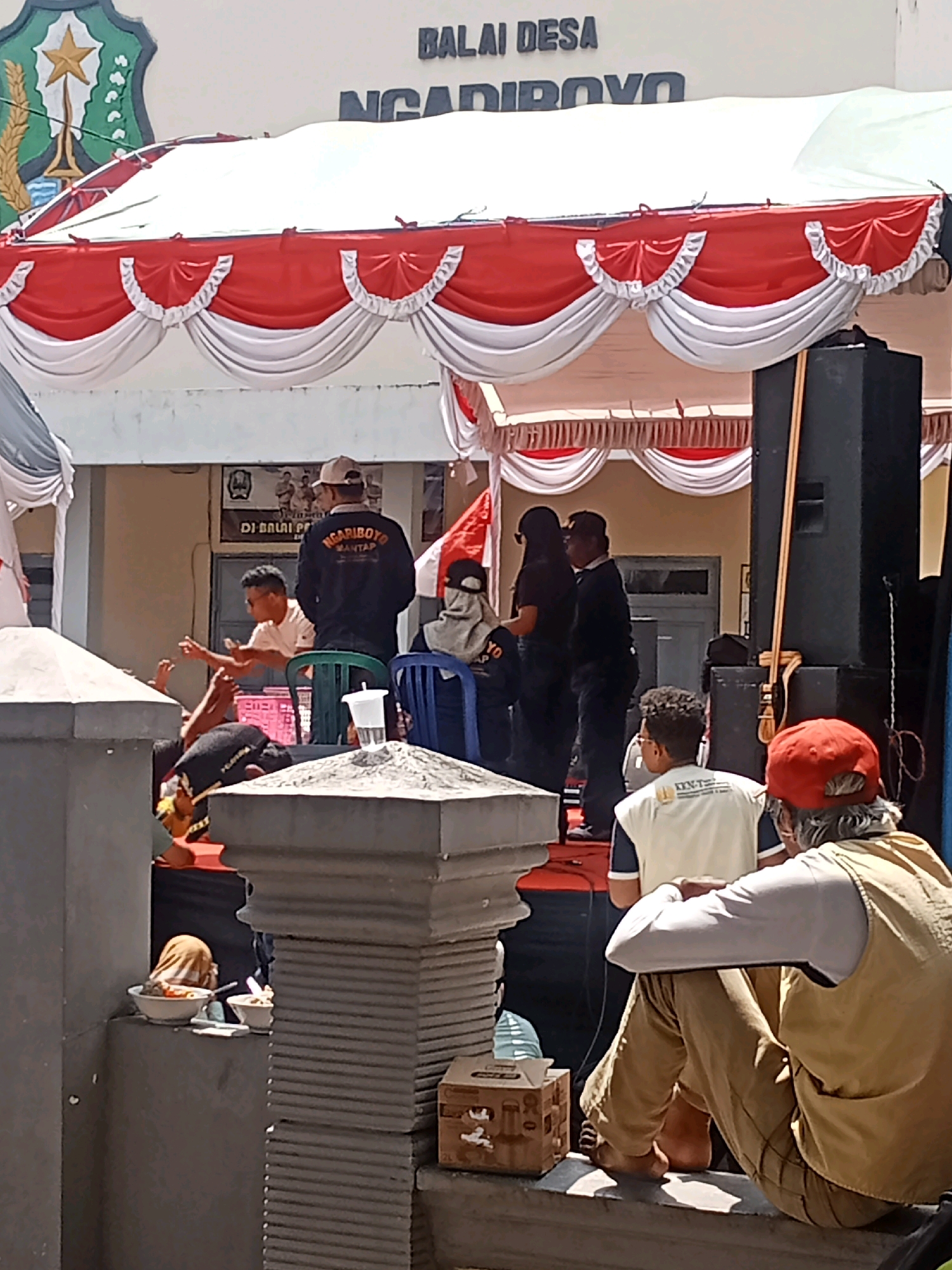 The image captures an outdoor event at the Ngariboyo Village Hall (Balai Desa Ngariboyo) in Ngariboyo, Indonesia. A stage is set up with red and white draped decorations. Several individuals are present, some on the stage and others in the foreground. On the stage, a group of people appear to be interacting, with one person in a black jacket with "NGARIBOYO MANTAP" written on it. A large black speaker system is visible to the right of the stage. In the foreground, a person wearing a red cap and a tan vest is seated, facing away from the camera, with their legs crossed. Another person in a tan vest is also visible. To the left of the image, two bowls of food are placed on a ledge. The overall scene suggests a community gathering or event.