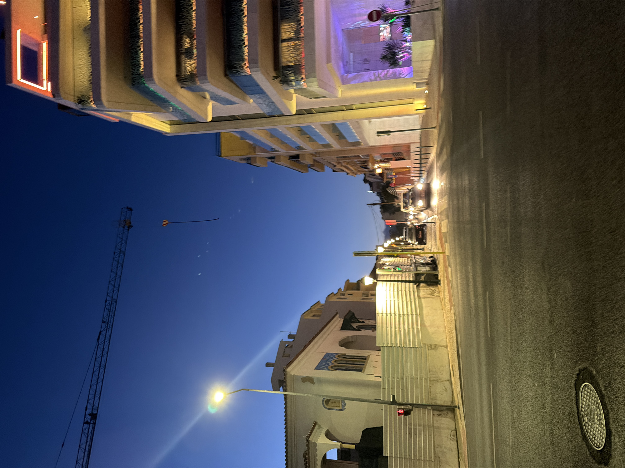 The image depicts a street scene in Cascais, Portugal, at dusk or early night. The perspective is from a street-level view looking up towards buildings and the sky, with the camera rotated 90 degrees counterclockwise.
On the left side of the frame, a tall construction crane with a long boom extends upwards into the deep blue twilight sky. A thin string or cable hangs from the crane boom, with a small, multi-colored kite attached to its end.
The buildings lining the street are varied. Some appear to be modern apartment or hotel buildings with balconies and illuminated windows, showcasing a mix of warm and cool lighting. Others are older, featuring tiled accents and arched window details, suggesting a blend of architectural styles. Construction fencing, made of corrugated metal, partially obscures some of the buildings.
Streetlights are illuminated, casting a warm glow on the asphalt road and sidewalks, highlighting the wet pavement, which suggests recent rain or damp conditions. Several cars are visible parked or driving along the street, their headlights and taillights adding to the nighttime ambiance. A traffic signal with a red light is also visible.
The overall mood is one of a quiet evening in a coastal town, with the transition from day to night creating a sense of calm and perhaps a touch of mystery. There is no visible text in the image.