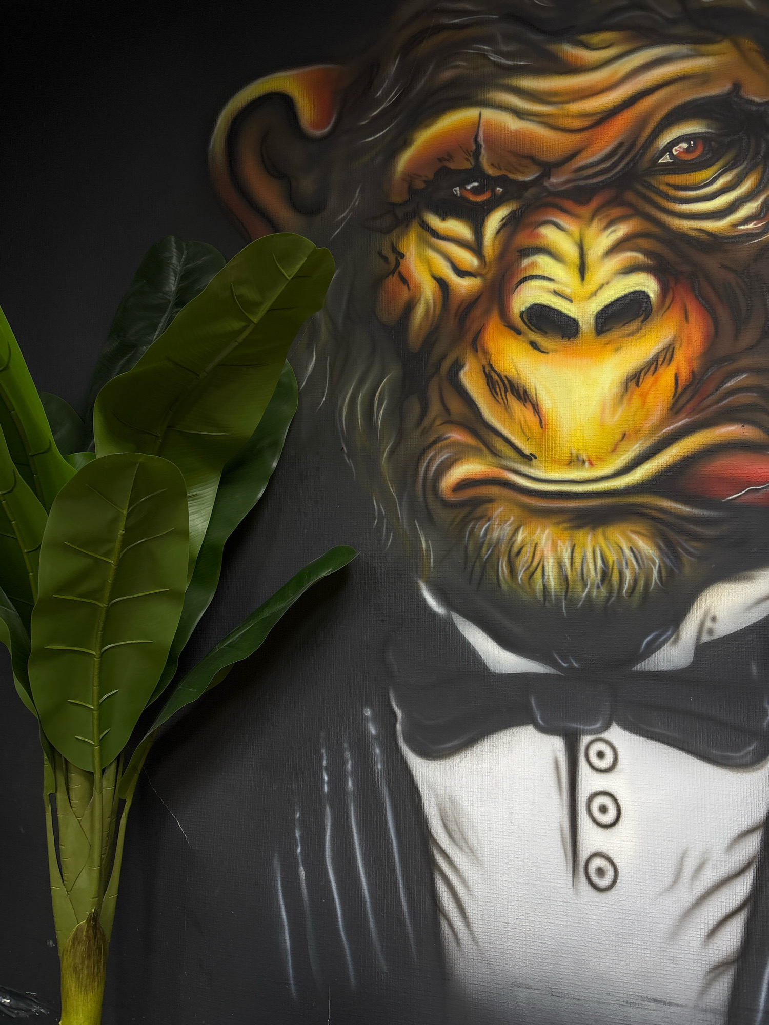 The image is a close-up, vertical shot of a painting depicting a chimpanzee wearing a tuxedo. The chimpanzee's face is rendered in warm tones of orange, yellow, and brown, with detailed fur texture and piercing eyes. A cigar is visible in the chimpanzee's mouth. In the foreground, to the left of the painting, a cluster of large, vibrant green artificial leaves from a plant are prominently featured. The background of the painting is a dark, textured surface, possibly canvas. The overall aesthetic suggests an artistic display. There is no discernible connection to the city of Ufa, Russia, based on the visual information present in the image.