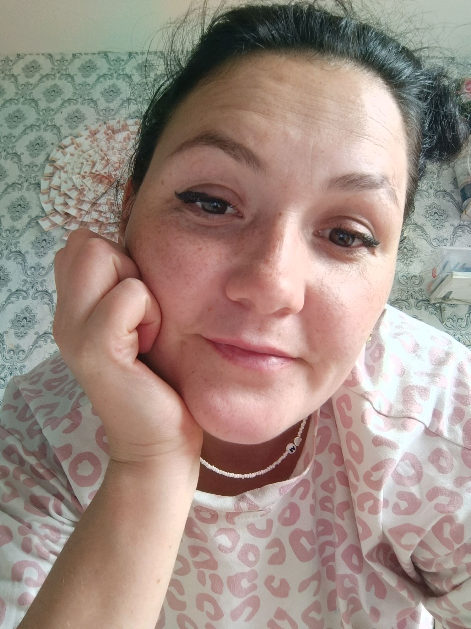A close-up, high-angle, outdoor selfie shows a Caucasian woman with dark hair pulled up in a bun, resting her chin on her hand. She has freckles on her face and is wearing eye makeup and a light-colored shirt with a pink leopard print pattern and a beaded necklace with the letter "M" on it. The background consists of patterned wallpaper. The image does not contain any information that would indicate the location is in Chaikovsky, Russia.