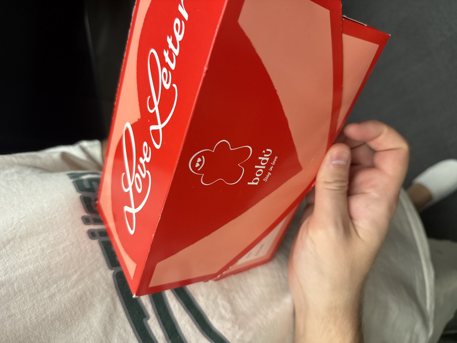 A partially visible individual is holding two stacked red rectangular boxes, presumably containing pastries or donuts, in an indoor setting. The top box is prominently branded with "Love Letter" in white script over a light red heart graphic on its lid. The front face of this box displays a white outlined gingerbread-man-like figure with heart eyes, the brand name "boldú," and the slogan "Stay in love." The individual's right hand grasps the boxes. Their torso, wearing a light-colored t-shirt with a dark horizontal stripe or graphic, is visible in the upper left, while their left leg and a white sock are visible in the lower left. The background is dark and out of focus, with a discernible dark floor. The "boldú" branding indicates the products are from a Boldú bakery/donut shop located in Barcelona, Spain.