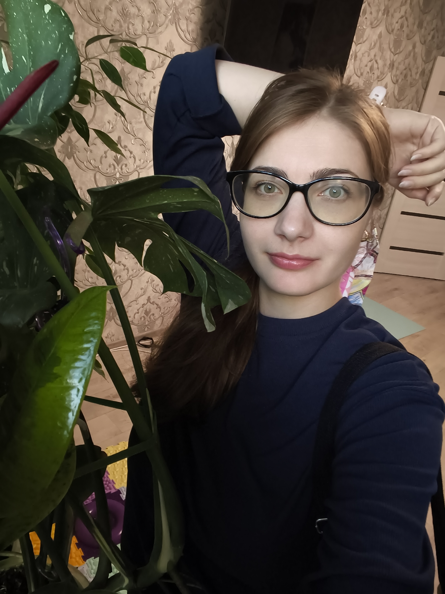 The image is a selfie of a young woman with reddish-brown hair and glasses, taken indoors. She is wearing a dark blue long-sleeved shirt and has her left arm behind her head. Her expression is a slight smile. To her left is a large green houseplant with large, split leaves and a reddish-purple spadix. The background shows a patterned wallpaper, a wooden floor, and part of a doorway with a white door. A colorful interlocking floor mat is partially visible on the floor. The lighting suggests it is daytime, and the indoor setting indicates she is at home or in a similar private space. The woman's pose and slight smile suggest a casual and relaxed mood. There is no visible text in the image.