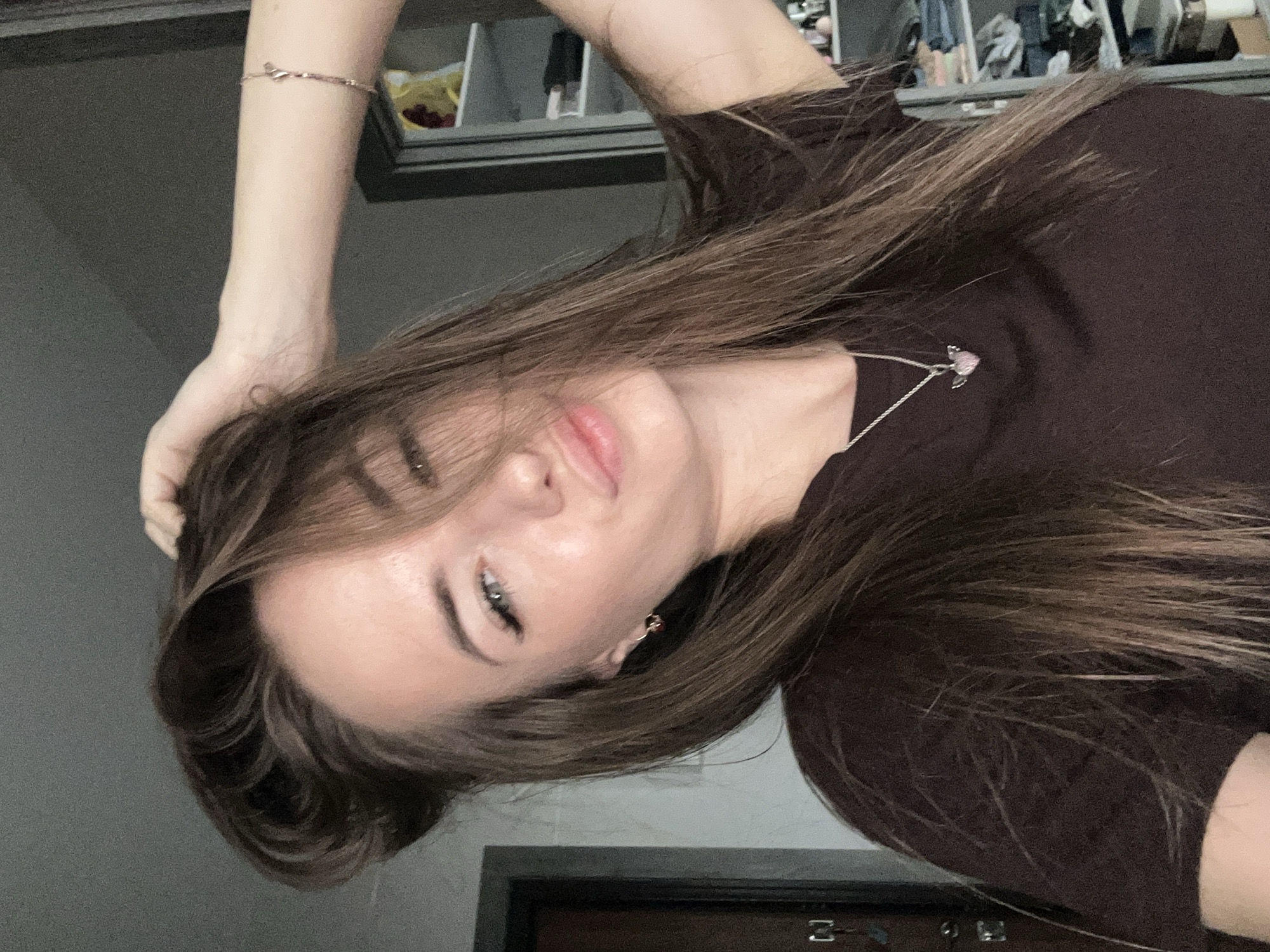 An upper body, medium shot captures a young adult woman with long, dark brown hair, positioned slightly off-center to the right. She is looking towards the camera with a playful expression, her lips pursed. Her right hand is raised, fingers tucked into her hair. She is wearing a dark brown t-shirt with a delicate silver necklace featuring a small pink heart pendant with wings. A thin gold bracelet adorns her right wrist. The background is dimly lit, showing a grey wall on the left, a door with visible hardware, and a closet or shelving unit on the right filled with various items. The lighting appears to be natural, coming from the front and slightly from the right. The woman's skin is smooth and her makeup includes eyeliner and mascara.
