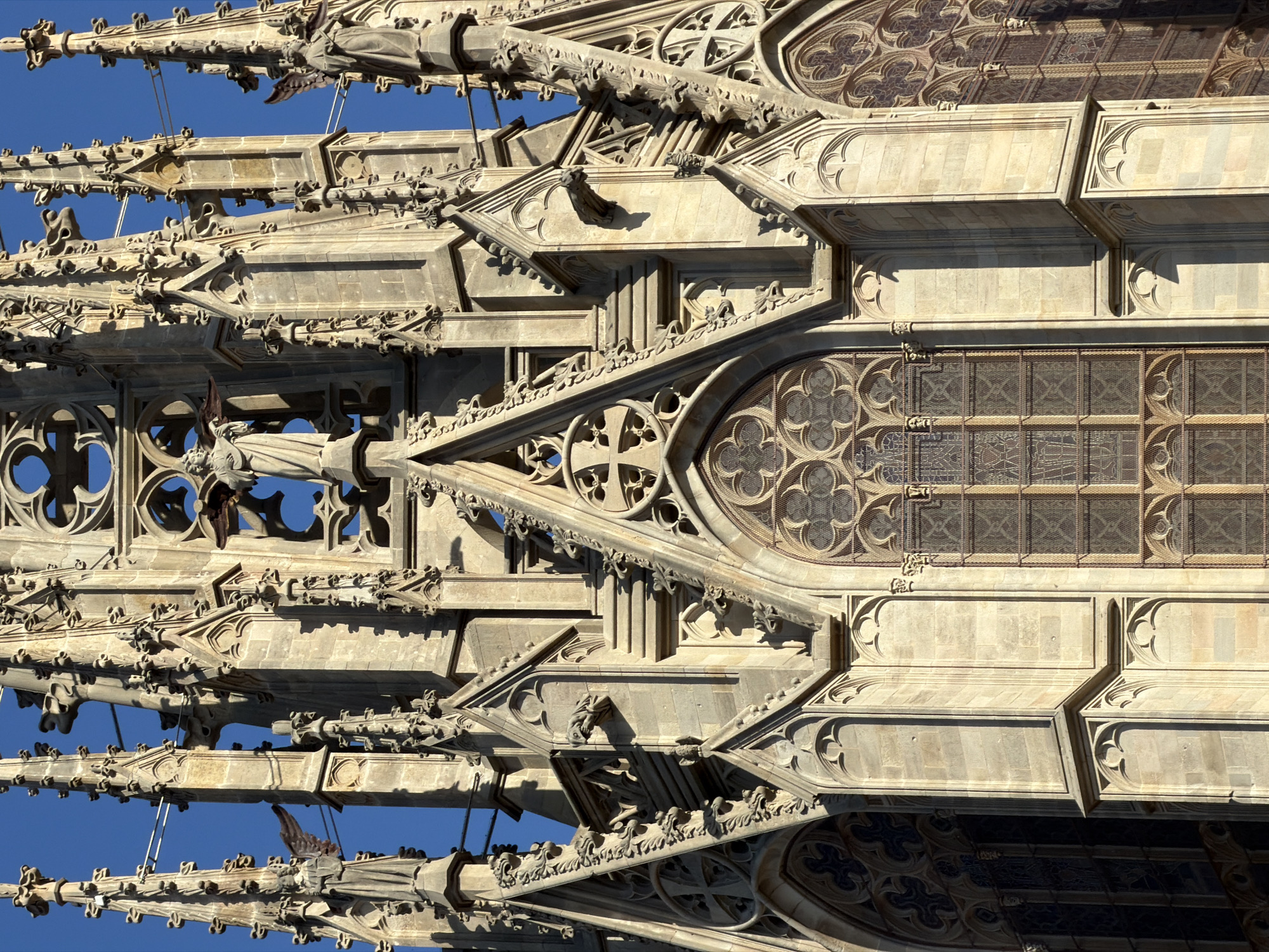 This media file shows an extreme close-up, low-angle shot of the intricate Gothic architecture of a cathedral's exterior, likely Barcelona Cathedral in Spain, against a clear blue sky. The composition is dominated by the detailed stone carvings of flying buttresses, spires, gargoyles, and ornate window tracery. Several small gargoyles, designed to channel rainwater away from the building, are visible perched on ledges and protruding from corners, adding a touch of the grotesque to the otherwise majestic stonework.
The architectural elements are carved from light-colored stone, suggesting it is either sandstone or limestone, and they are heavily ornamented with decorative moldings, pinnacles, and quatrefoil and trefoil motifs characteristic of Gothic design. Sunlight casts strong shadows across the stonework, highlighting the three-dimensional complexity of the structure and indicating that the photograph was taken during the daytime, likely in the late morning or early afternoon given the angle of the shadows. The bright blue sky without any clouds suggests clear, fair weather.
The image's perspective creates a sense of overwhelming scale and grandeur, drawing the viewer's eye upwards along the ascending lines of the architecture. The overall impression is one of ancient craftsmanship, religious devotion, and the enduring beauty of Gothic cathedrals. No people or text are visible in the image.