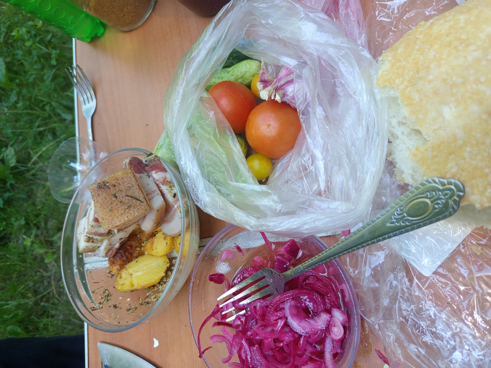 The image depicts a close-up, overhead view of a wooden table set with food items, likely outdoors due to the visible greenery in the background and the casual arrangement of the meal. In the foreground, a torn piece of bread rests on a clear plastic bag. To the left, a glass bowl contains thinly sliced red onions, with a silver fork resting inside. Behind this bowl, another glass bowl holds pieces of cooked potatoes and cured meat with herbs. Further back, a clear plastic bag is filled with fresh vegetables including tomatoes, cucumbers, and garlic. A green bottle and a glass are partially visible in the upper right of the frame. The overall scene suggests a picnic or an outdoor meal. The location is identified as Giaginskaya, Russia.