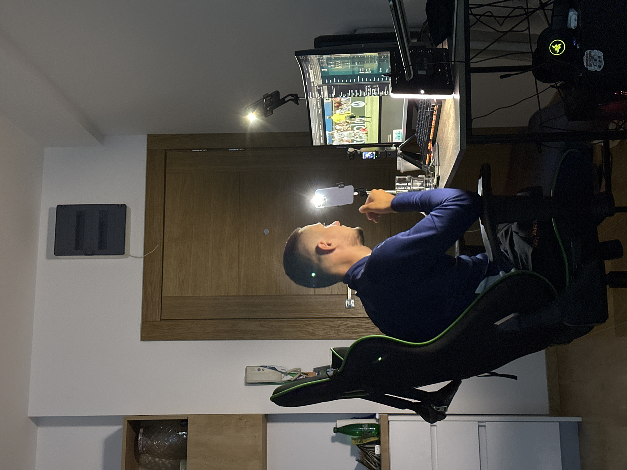 In the city of Donji Pobori, Montenegro, a young man is seated in a black and green gaming chair, facing a desk with a computer setup. He is wearing a dark blue long-sleeved shirt and appears to be looking intently at the computer monitor. On the desk, there is a computer monitor displaying a sports game, a keyboard, a glass of water, and various electronic devices including a smartphone mounted on a tripod and a ring light. Behind the man, a wooden door is visible, and to the left, a shelf with a decorative jar. The room is dimly lit, with the primary light sources being the computer monitor and the ring light. The overall environment suggests a personal workspace or entertainment area.