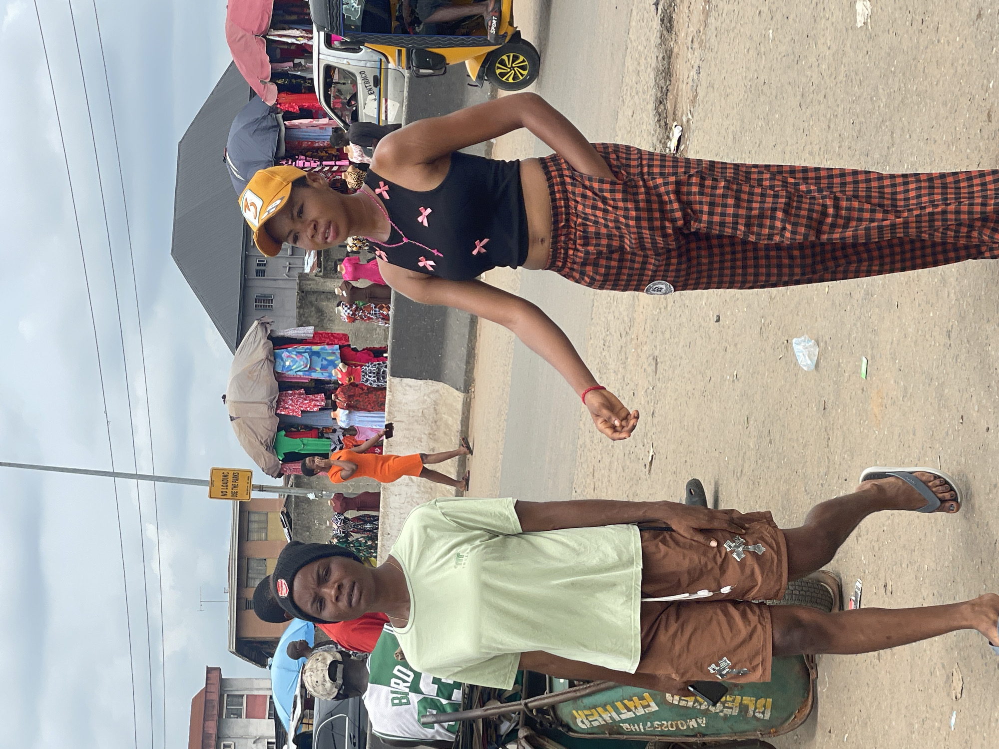 The image shows two individuals posing on a street in Onitsha, Nigeria, amidst a bustling market scene. One individual, wearing a yellow baseball cap, a black crop top with pink bow accents, and orange plaid pants, stands on the right side of the image. The other individual, wearing a black beanie, a light green t-shirt, and brown shorts, stands on the left. In the background, there are vendors selling clothing, a yellow auto rickshaw, and pedestrian traffic. A traffic sign is visible on a pole nearby. The surrounding environment depicts a typical urban market scene with various commercial activities and urban infrastructure.