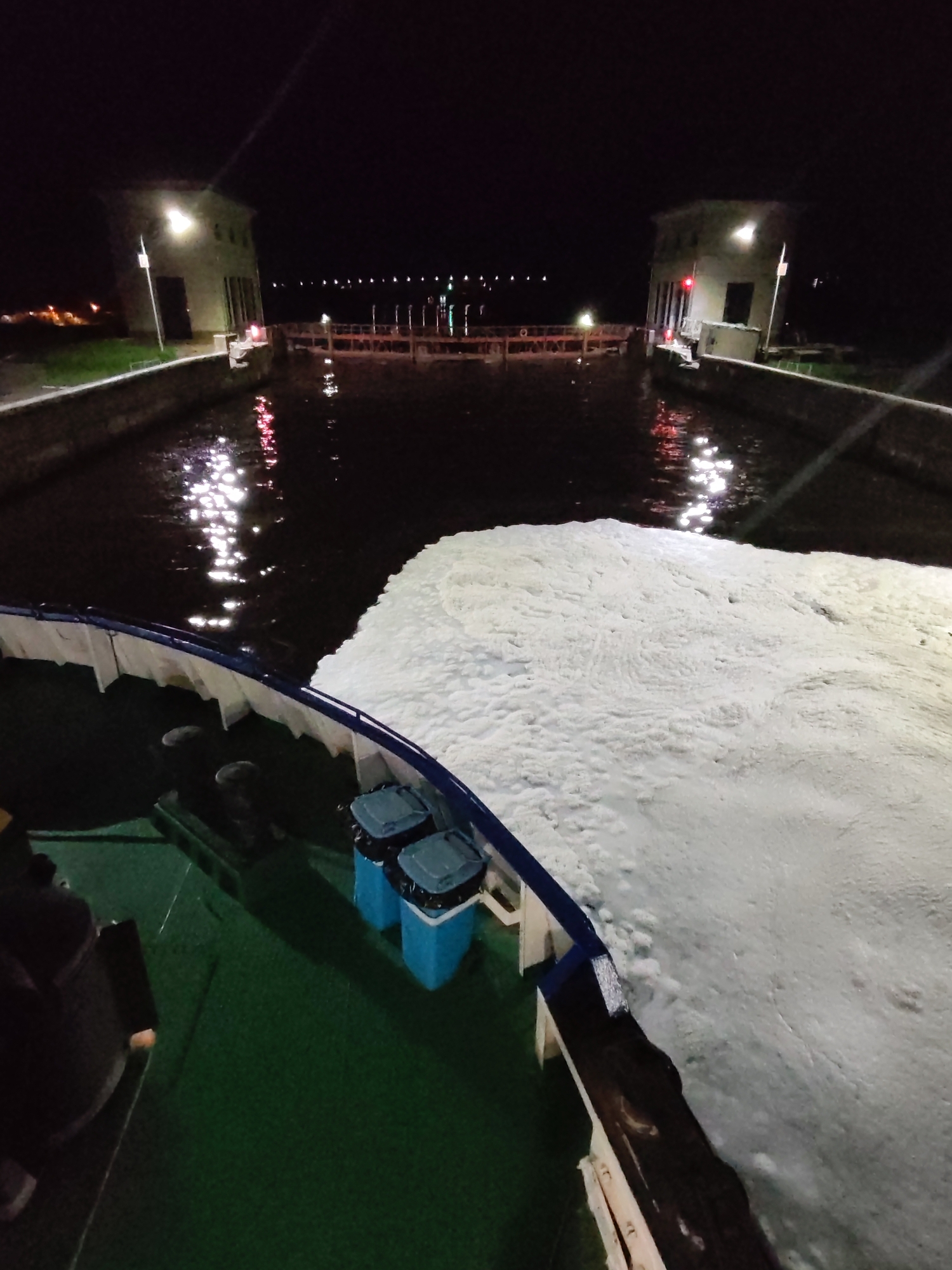 The image captures a night scene in Dubna, Russia, showing a canal lock filled with white foam. The view is from a boat, with the vessel's green deck and blue trash cans visible in the foreground. The white foam covers a significant portion of the right side of the image, extending from the boat toward the canal. On either side of the canal are two lock control buildings, lit by lamps. In the distance, a series of lights line a bridge or structure over the water. The water reflects the lights, adding to the overall brightness of the dark environment.