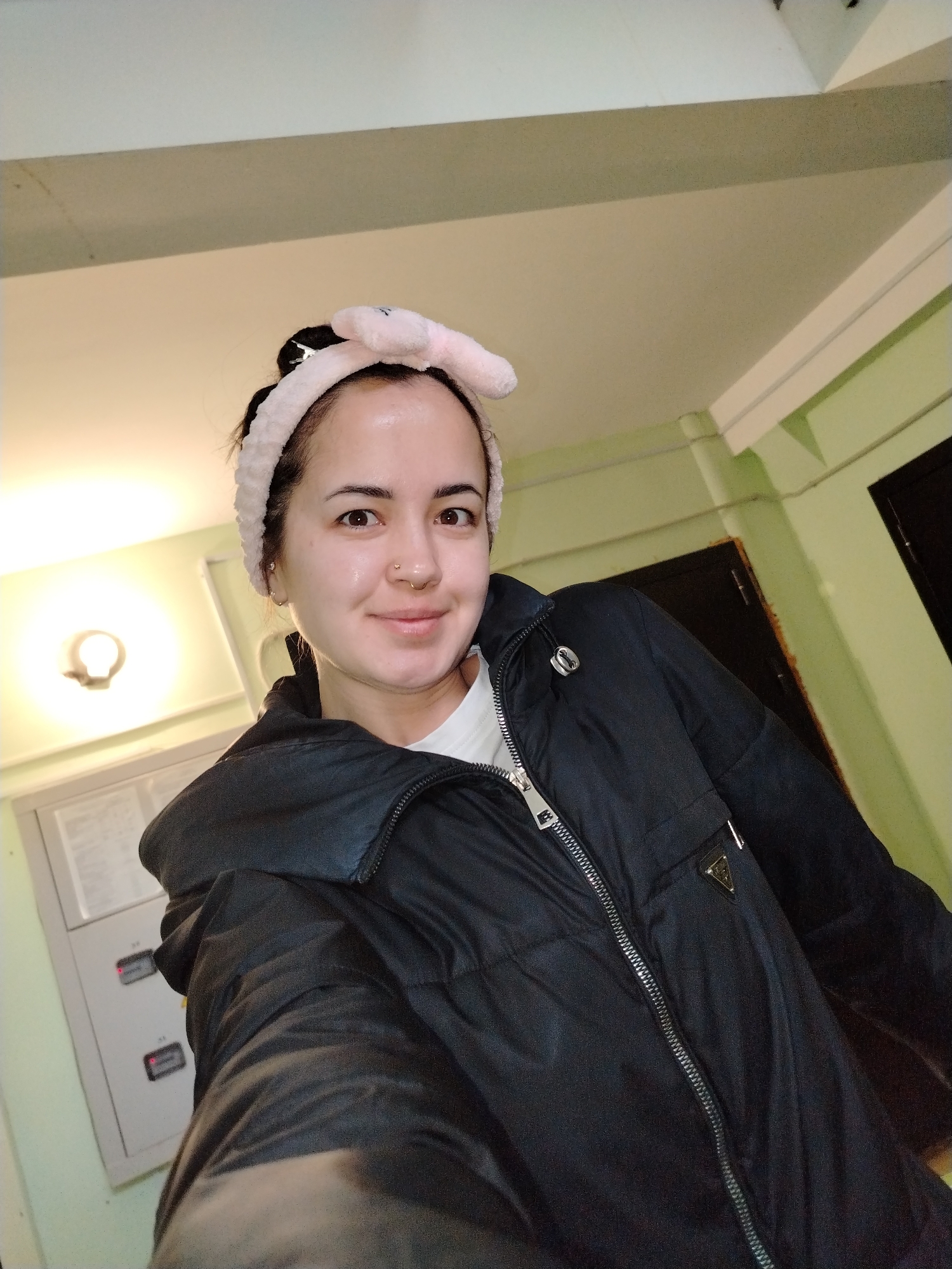 A woman is depicted in a close-up, indoor selfie, looking directly at the camera with a faint smile. She has dark hair pulled back with a soft, pink, bunny-ear headband. She wears a silver nose ring and an earring in her left ear. Her skin appears moisturized and glowing. She is dressed in a black puffer jacket with a prominent silver zipper and what appears to be a white t-shirt underneath. On the upper left arm of her jacket, a shiny, black triangular emblem is visible.
The setting appears to be an interior space, possibly a hallway or utility area, with a beige ceiling and upper wall sections, contrasted by light green lower walls and structural beams. On the left side of the frame, a wall-mounted light fixture emits a warm glow. Below it, a grey electrical panel is visible, featuring two rectangular indicators glowing red and some faint, illegible text or numbers. A dark door or entryway is partially visible in the upper right background. The lighting is entirely artificial, suggesting the photo was taken indoors, likely in the evening or at night in Noviy, Russia.