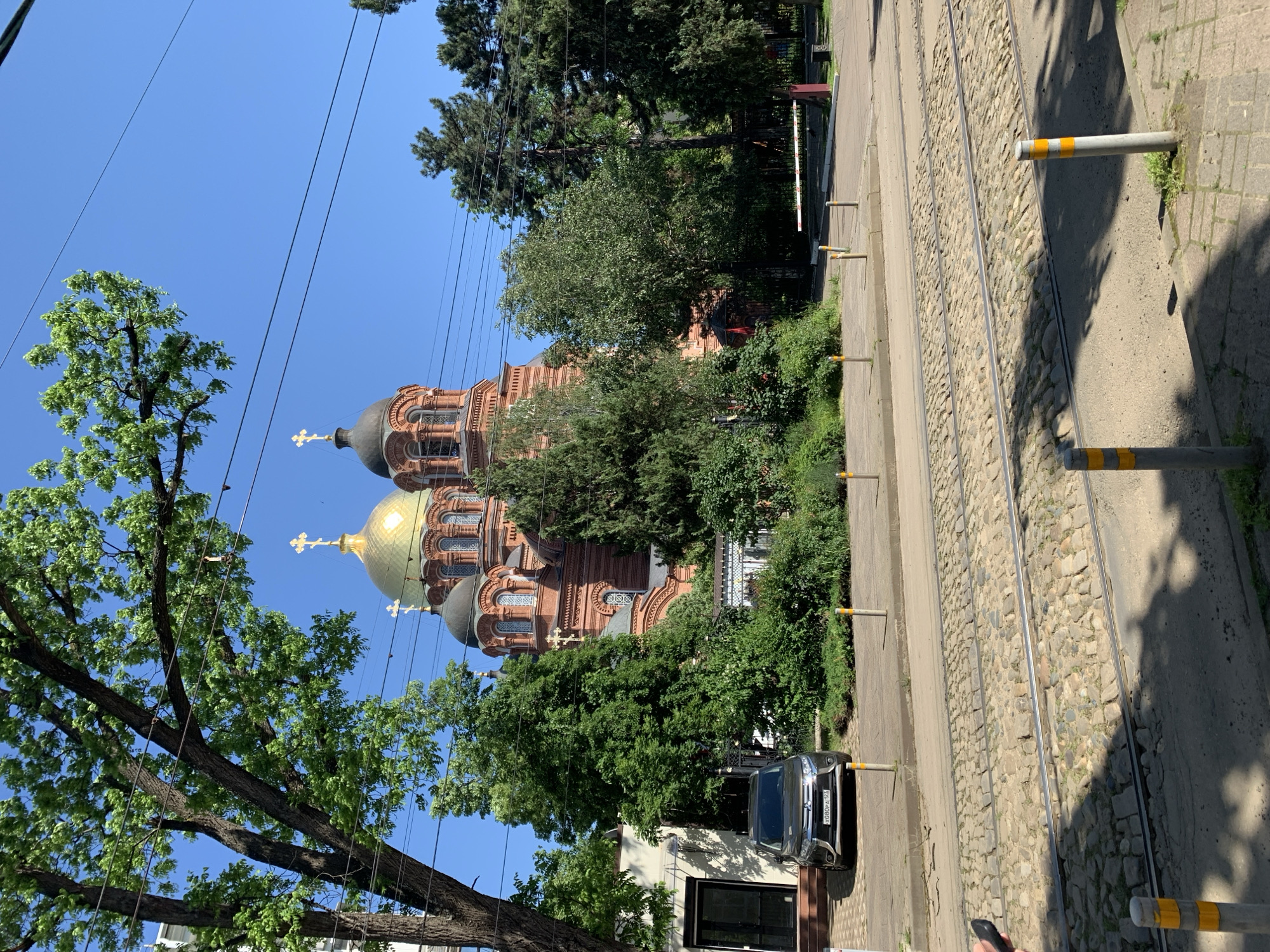 The image depicts a Russian Orthodox church partially obscured by trees, a street with tram tracks made of cobblestones, and a car parked on the street. The church has a golden dome and a darker dome with crosses on top. There are overhead wires in the sky. The location is not identifiable from the image.