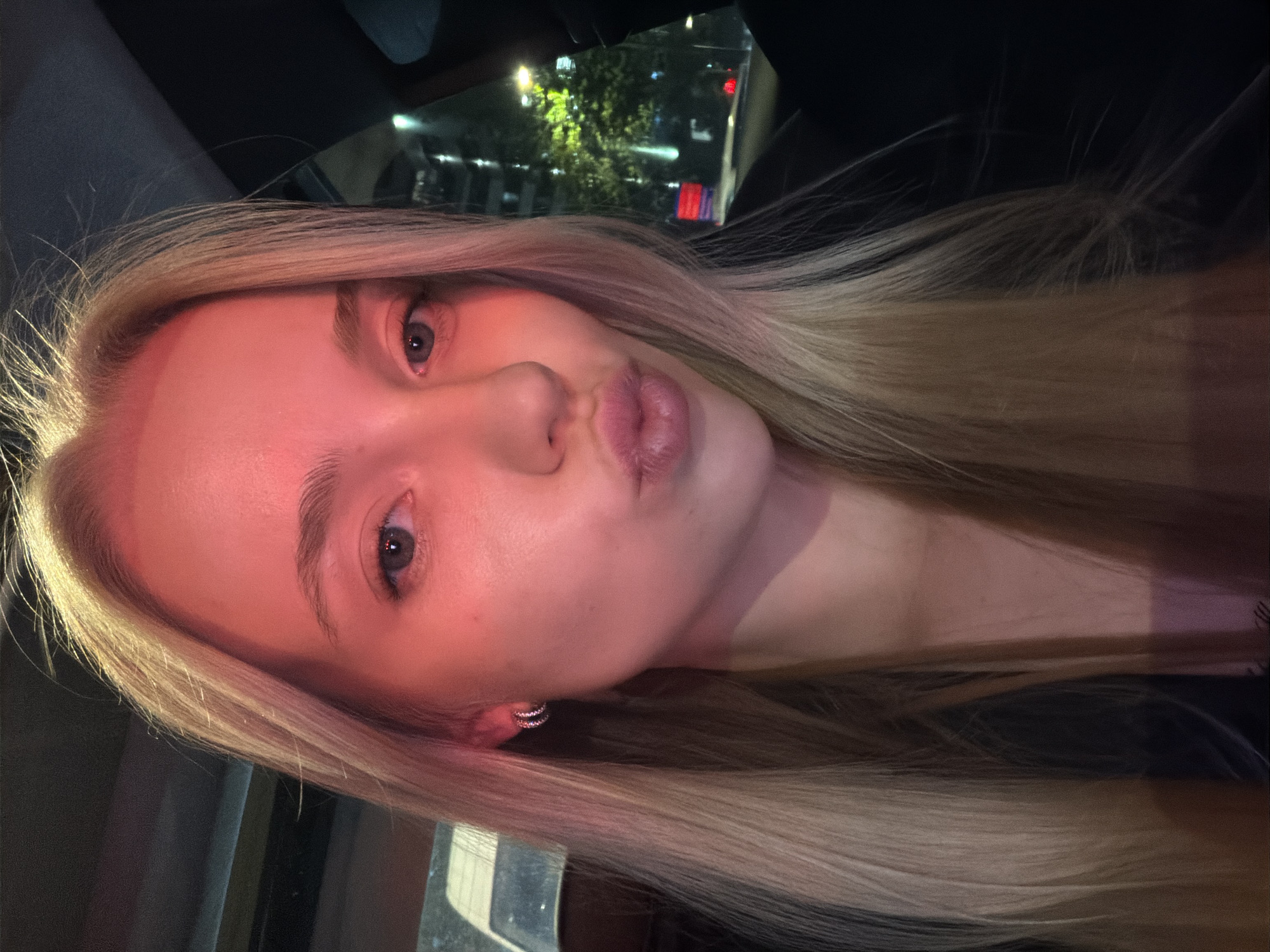 A person with light blonde hair is facing the camera with pursed lips. The person appears to be in a car at night, as reflections of city lights and buildings are visible through the car window. The face is illuminated with a reddish tone. The location in the city is unknown.