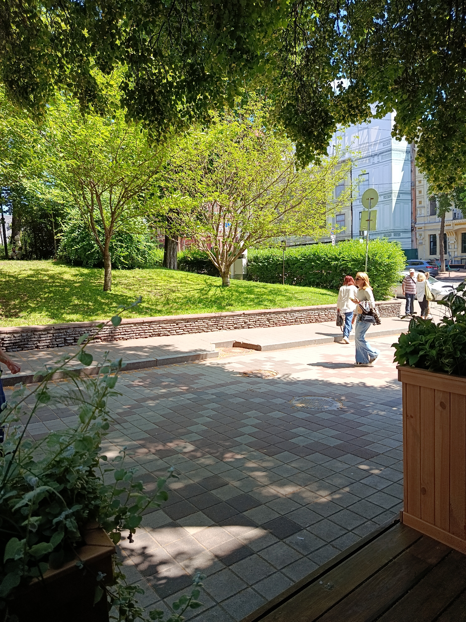 This image captures a sunny street scene in Kyiv, Ukraine, likely on a warm day. The foreground features a textured pavement of grey and patterned square tiles, with dappled sunlight and shadows cast by overhead foliage. To the right, a wooden planter box contains lush green plants.
In the middle ground, a low, decorative stone wall borders a grassy embankment. Several people are walking along the sidewalk to the left of the wall. One woman in the foreground, wearing blue jeans and a light-colored top, is walking away from the camera with a purse slung over her shoulder. Behind her, another woman in a white top and dark pants also walks away. Further in the background, two more individuals, a man and a woman, are walking in the same direction.
The background reveals urban architecture, including a building with white and light blue facades adorned with decorative moldings. Street signs are also visible. Trees with vibrant green leaves provide a leafy canopy over the scene, creating a pleasant, shaded atmosphere. The overall impression is of a public urban space, possibly a park or a street with a green zone, being enjoyed by pedestrians on a bright day.