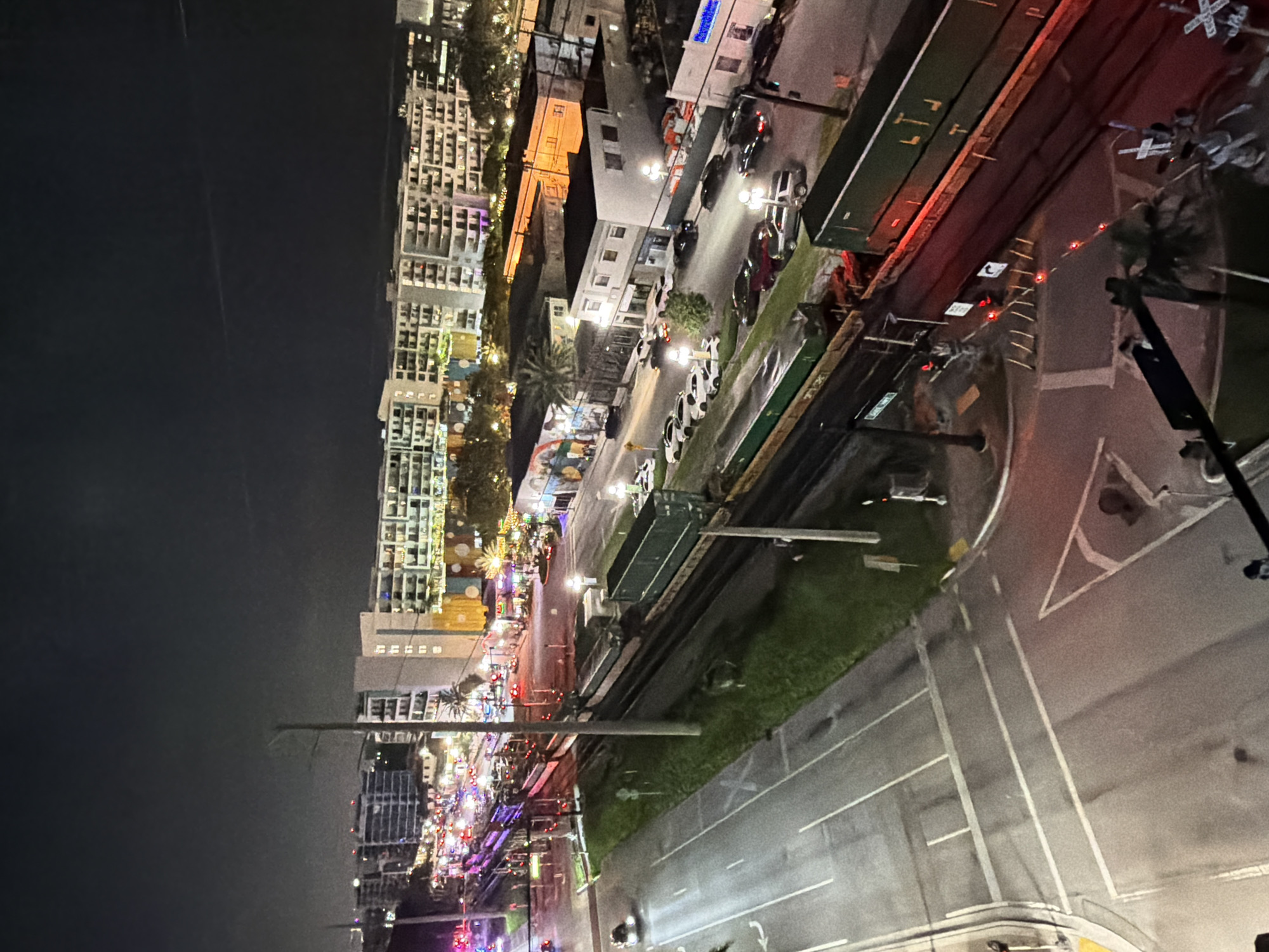 This is an aerial, nighttime photograph of a city scene in Hollywood, Florida, USA. In the foreground, a train with green cargo containers is stopped on the tracks. The train crosses a road where traffic lights are visible, and other vehicles are stopped or moving. A street with parked cars and buildings lines the opposite side of the train tracks. Further in the background, a row of multi-story buildings, likely apartment complexes, are illuminated, with some displaying colorful murals on their lower levels. The sky is dark and cloudy, with a faint light suggesting urban illumination.