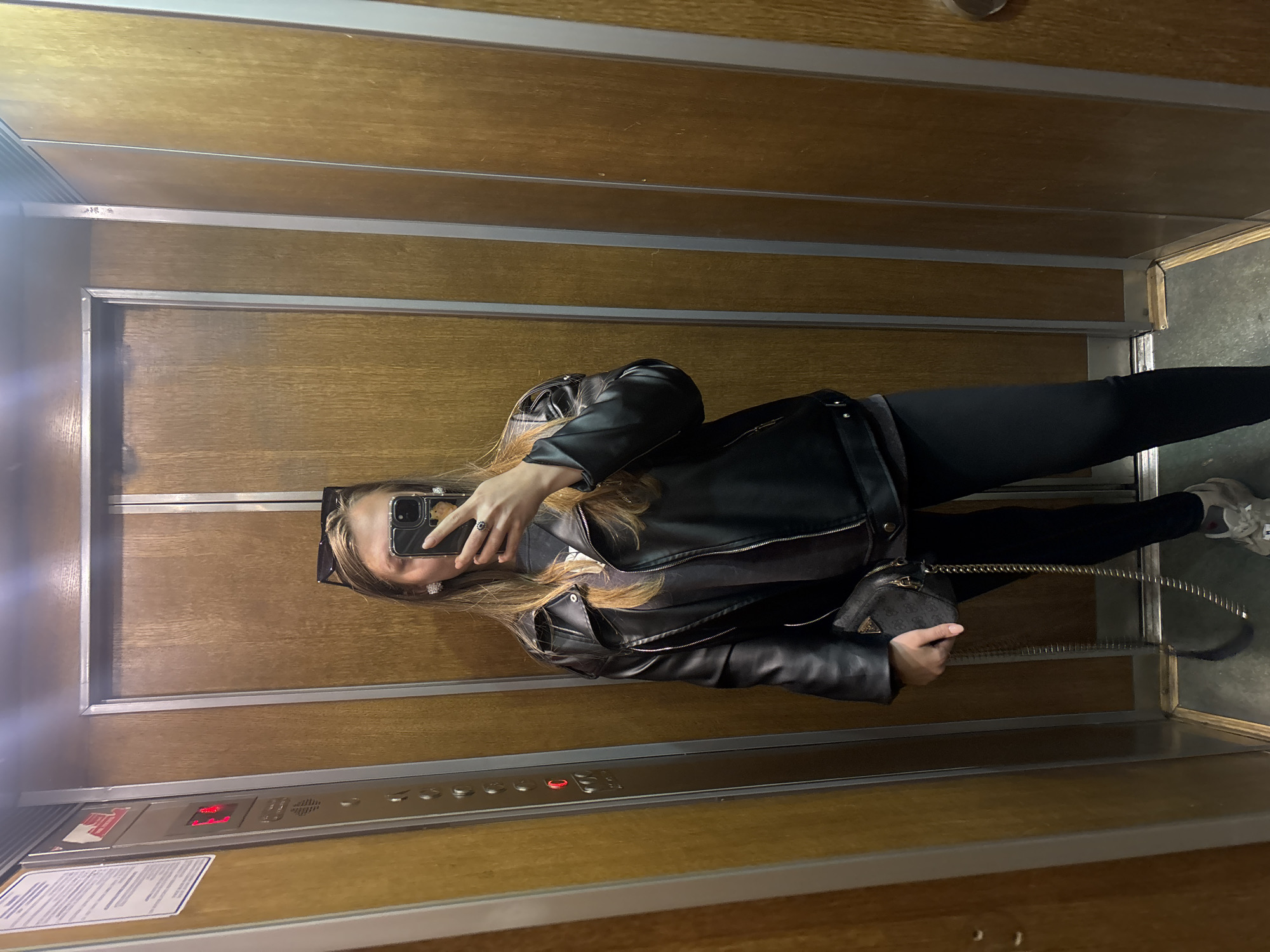 The image shows a woman in an elevator, taking a selfie in what appears to be a dated lift. She is wearing black leggings, a black leather jacket, and sneakers, with a small purse in her hand. The elevator is lined with wood paneling and metal trim, with a control panel visible near the bottom of the frame. The current floor is illuminated in red on the panel. The location is not apparent from the image; therefore, the city of Kharkiv, Ukraine, cannot be verified based on visual evidence alone.