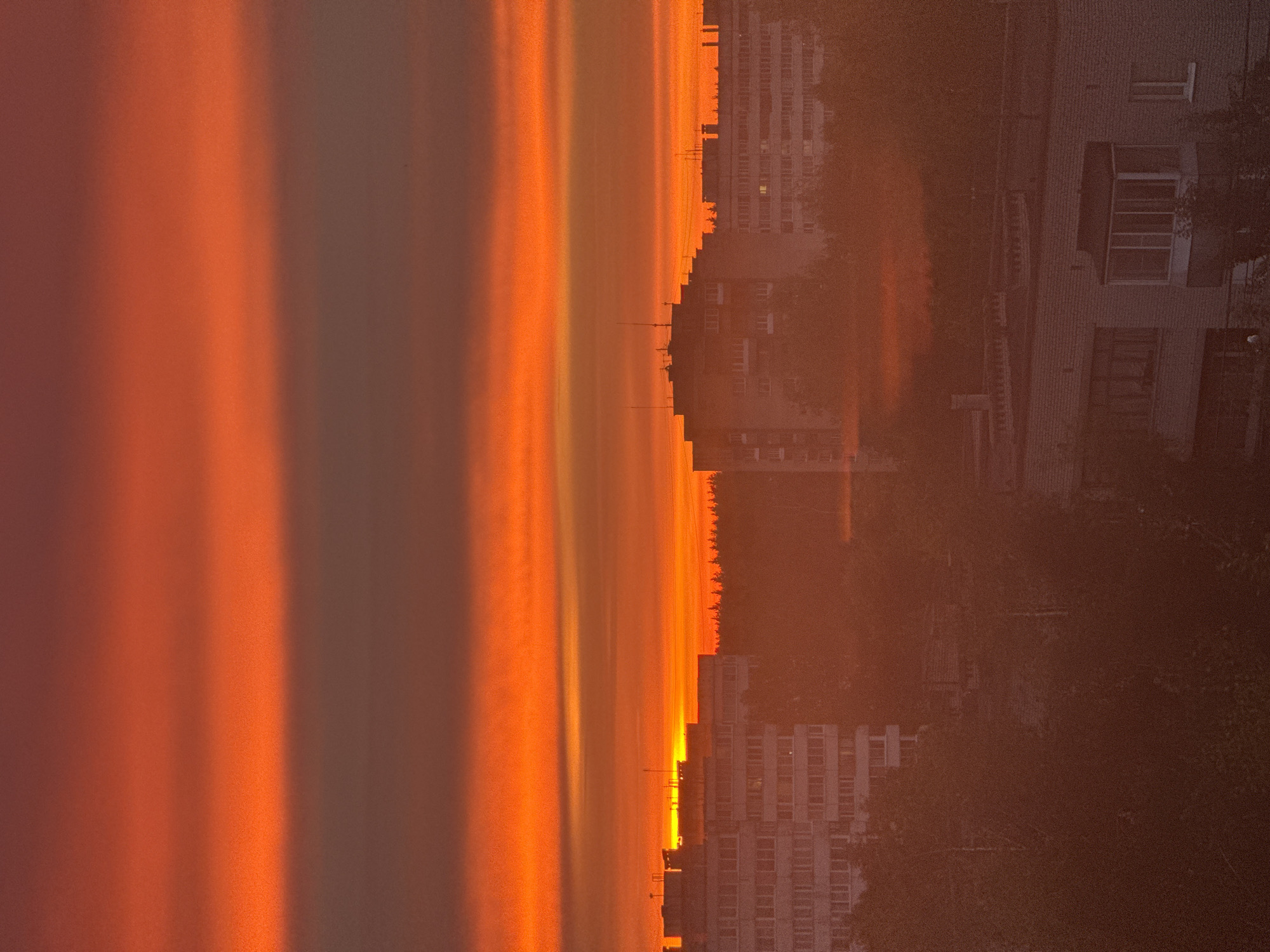 The image captures a wide shot of a city at dawn. The sky is dominated by vibrant hues of orange and red, indicating the sun is either rising or setting. The sky is layered with distinct bands of color, from bright orange near the horizon to darker reds and purples higher up. Silhouetted against this dramatic sky are several multi-story residential buildings, appearing as concrete structures with rows of windows. Some buildings are taller and more modern, while others seem older and lower. Trees and foliage are visible in the foreground and between buildings, adding a touch of nature to the urban landscape. The lighting suggests it is early morning or late evening, creating a hazy, atmospheric effect that softens the details of the city. The scene is set in Sosnoviy Bor, Russia, a location known for its nuclear power plant.