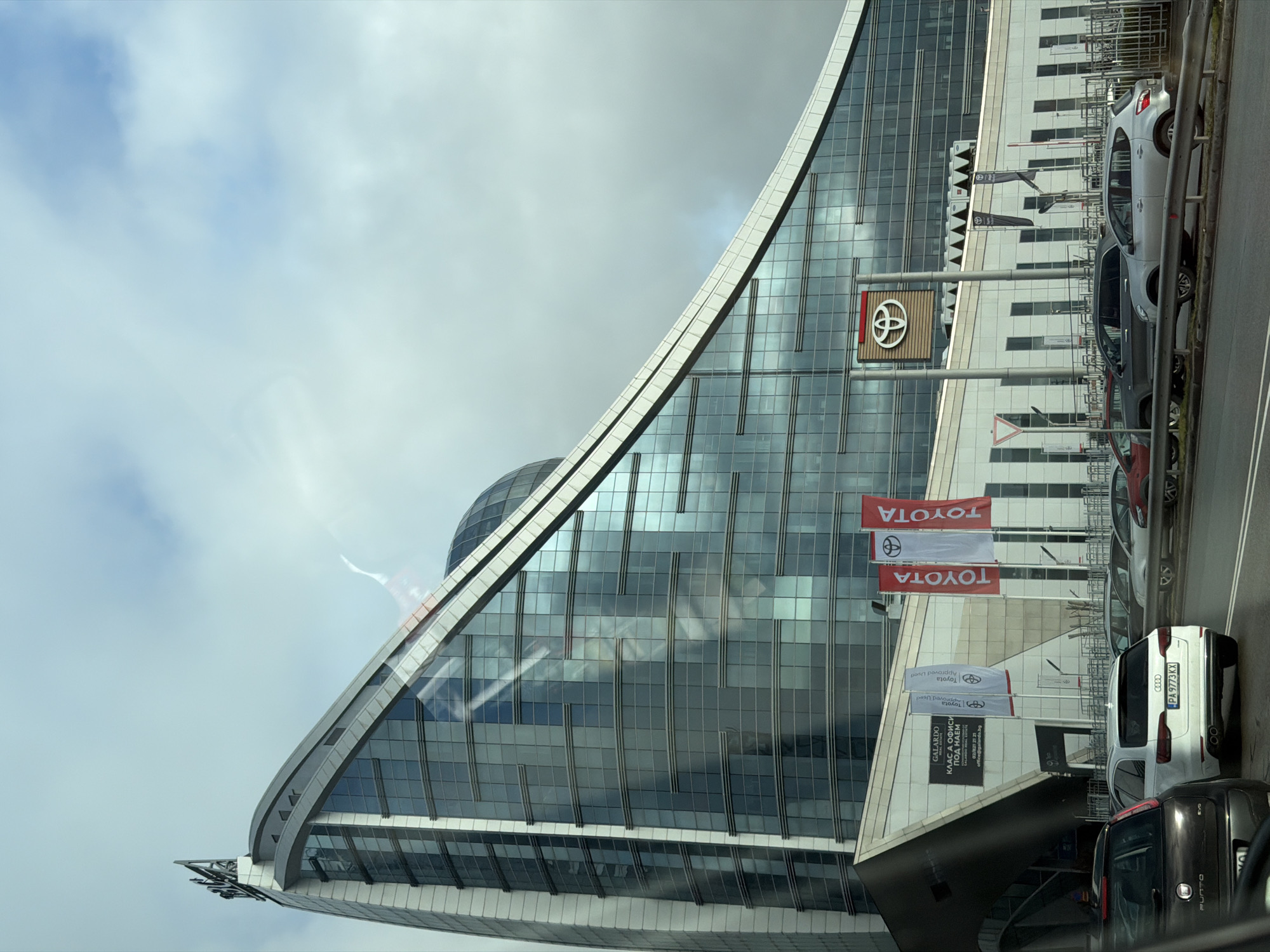 The image captures the exterior of a modern, triangular-shaped building with a domed structure partially visible behind it. This is the Sofia Airport Center, a business complex located in Sofia, Bulgaria. The building is predominantly glass with a grid-like pattern. In the foreground, there is a Toyota dealership, identifiable by the large Toyota logo and several vertical flags displaying the Toyota brand. Several cars are parked in front of the dealership. The sky is overcast, and there's a hint of contrail visible in the upper-left portion of the photo. A vehicle's dashboard is slightly visible at the bottom, suggesting the photo was taken from inside a car. Vehicle with number PA 9773 KX is partially visible.