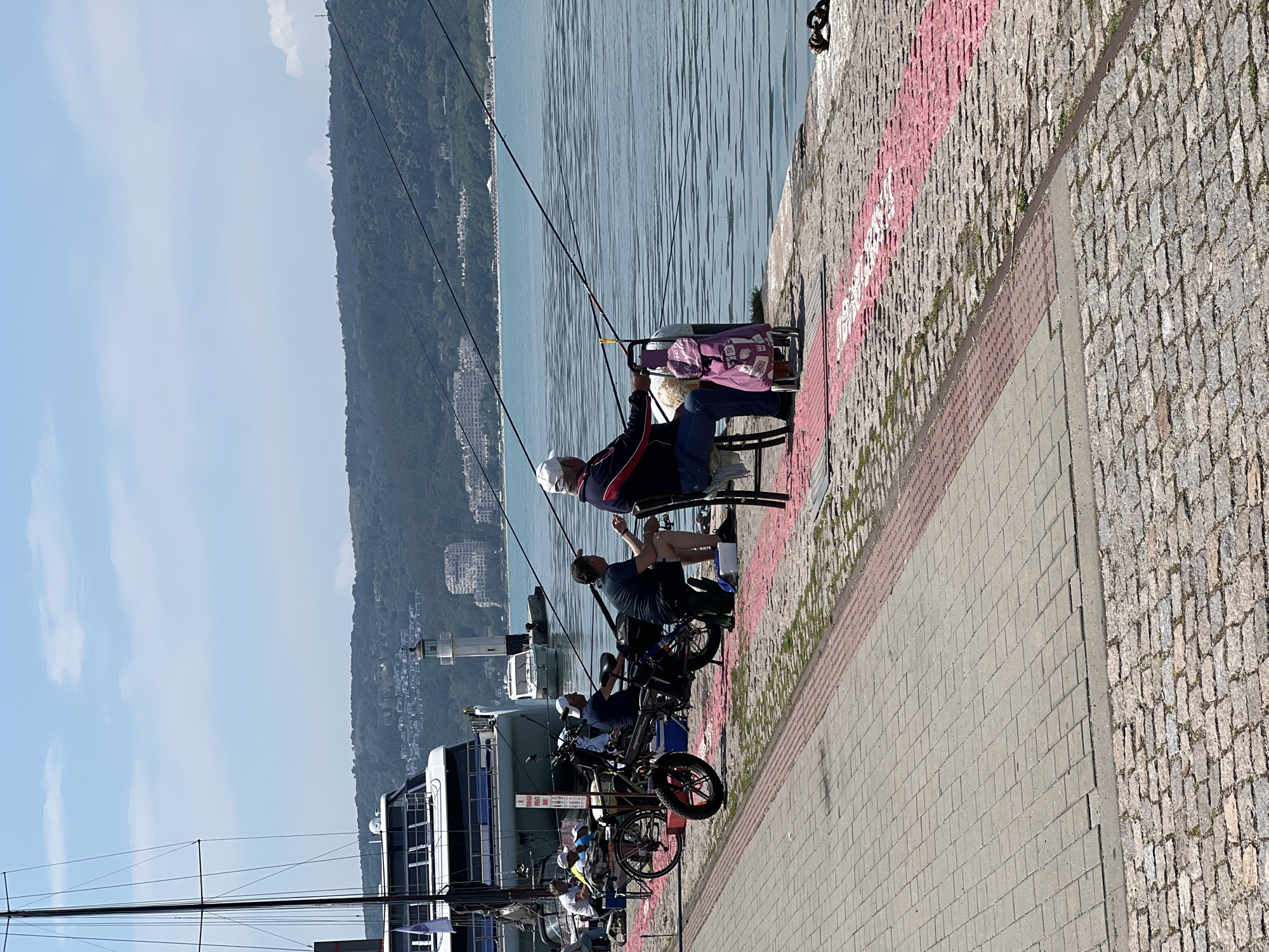 The image captures a vibrant waterfront scene along a pier in Varna, Bulgaria, on a clear, sunny day. The perspective is taken from the paved promenade, looking across a body of water towards a distant, tree-covered hillside dotted with buildings.
In the foreground, several individuals are engaged in leisure activities. Two men are prominently seated on chairs at the edge of the pier, fishing. One man, wearing a white cap and a dark jacket with red and white accents, holds a fishing rod, with a purple tote bag featuring the word "STOP" beside him. Another man, wearing a blue shirt, is also seated, with a fishing rod extending into the water. Several other individuals are visible further down the pier, some also seated, suggesting a communal fishing or relaxation spot. Bicycles or e-bikes are parked nearby, indicating casual transport or recreation.
The immediate ground features a combination of rough cobblestones and smoother paving slabs, with faint, faded pink or red paint markings and illegible words on the cobblestone section. The calm, blue-green water of the sea or a large lake fills the mid-ground, reflecting the sunlight. A large boat or ship is docked alongside the pier, partially visible on the left. In the background, lush green hills rise under a bright blue sky with a few scattered white clouds. Residential buildings are interspersed among the trees on the distant slopes.
The overall atmosphere appears relaxed and peaceful, typical of a pleasant day spent by the water. The strong sunlight suggests it is mid-day, casting clear but not overly long shadows.